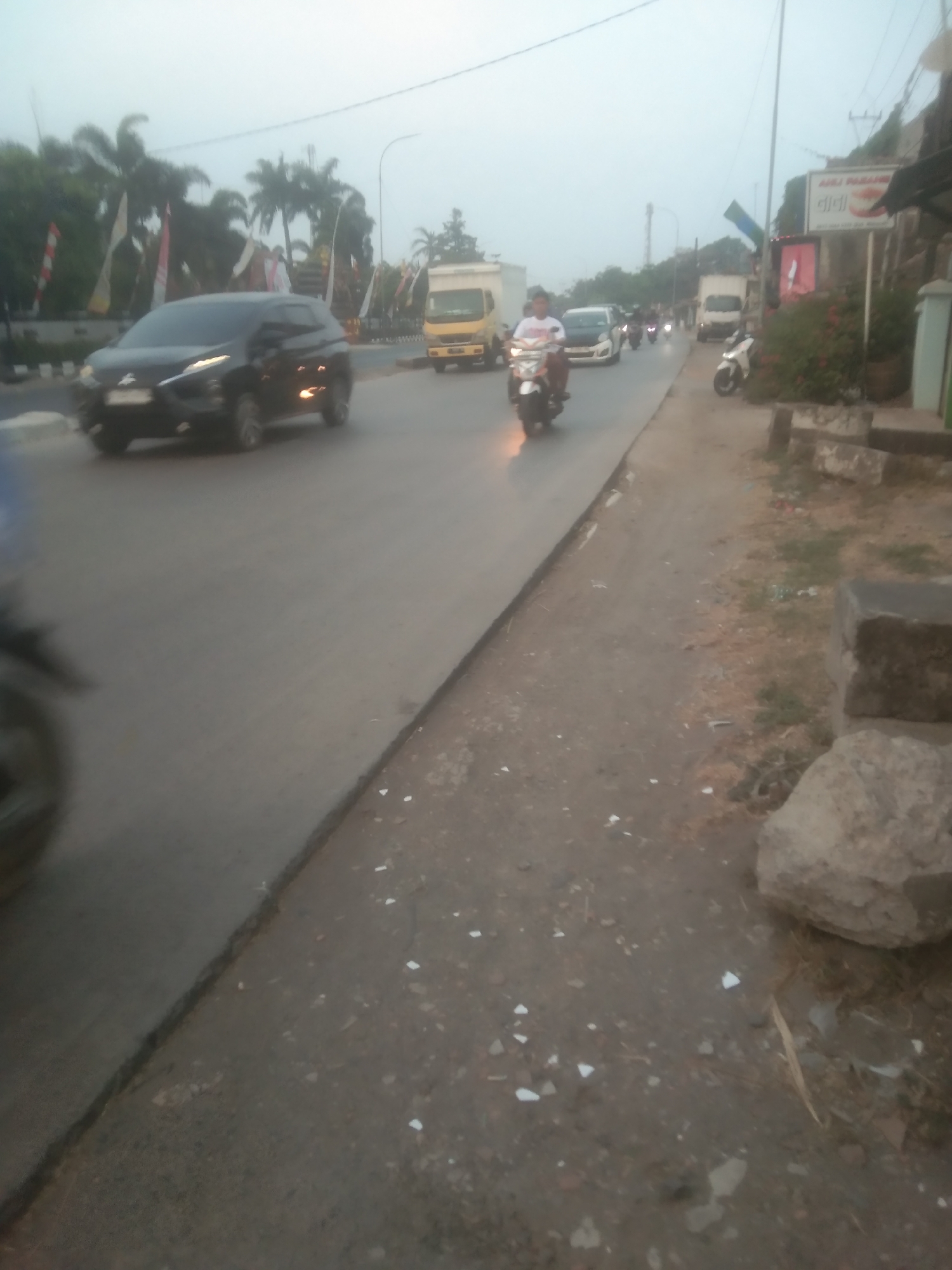 A motion-blurred, medium-shot, outdoor scene in Subang, Indonesia, depicts a busy street with multiple vehicles. In the foreground, a black SUV drives on the left side of the road. To the right, a sidewalk is visible, with a large rock and some debris scattered on the ground. In the background, several motorcycles and trucks are also present, along with a few palm trees and some buildings. The sky is overcast and the lighting is dim, suggesting it is either early morning or late afternoon. The overall impression is one of a typical day in an Indonesian city.
