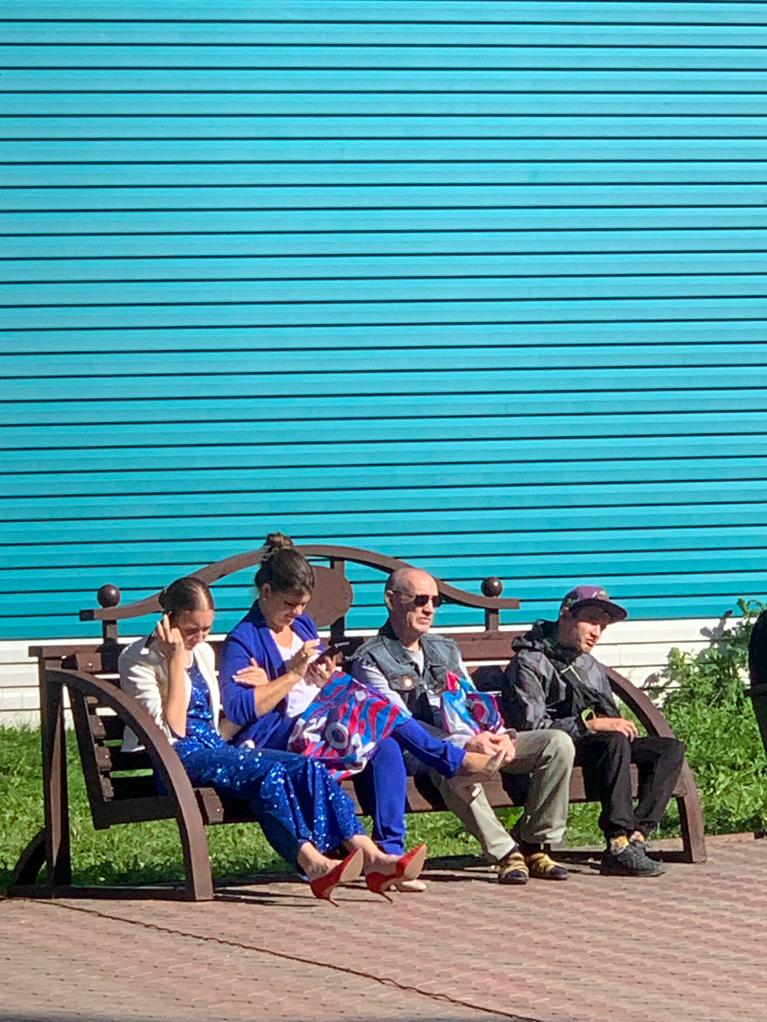 Four people are seated on a wooden bench against a bright turquoise, horizontally slatted wall. From left to right, the first person is a young woman wearing a white jacket over a blue sequined jumpsuit and red heels, with her hand to her ear. The second person is a woman in a blue blazer over a white top, looking at her phone. The third person is a man in sunglasses wearing a denim vest over a grey shirt and khaki pants. The fourth person is a young man wearing a dark hooded jacket, grey pants, and a purple baseball cap. The bench is on a brick pathway with green grass and foliage visible behind it. The scene appears to be outdoors on a sunny day.