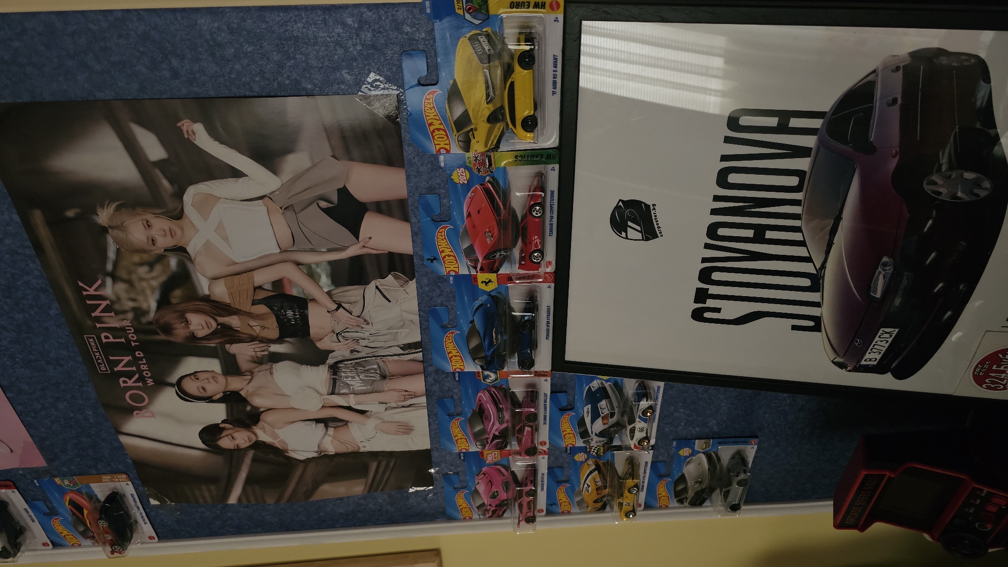 The image shows a wall decorated with various items. On the left is a poster of the K-pop group BLACKPINK, specifically promoting their "Born Pink" world tour. To the right of the poster are several Hot Wheels toy cars still in their packaging, arranged in a vertical row. Further to the right is a framed poster with the word "STOYANOVA" in large, bold letters next to a car. The car has the license plate "B 3773CK", which indicates it is registered in Bulgaria. The website "carposter.bg" is visible on the poster. The bottom right corner features a miniature arcade machine.
Based on the license plate and the website carposter.bg, it can be inferred that the location is in Bulgaria. However, without additional information from the image, pinpointing the exact location within Varna is not possible.