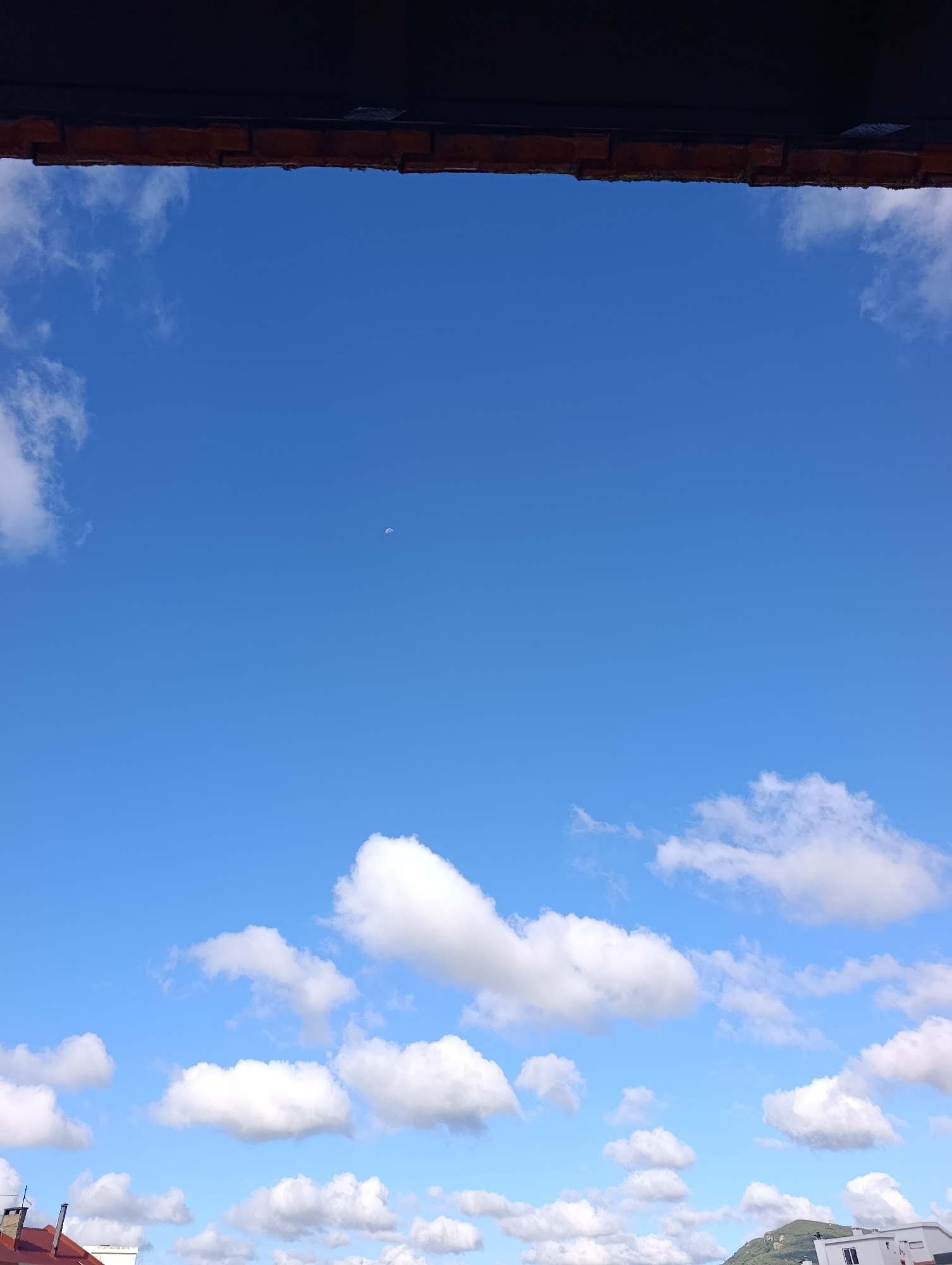The image depicts a clear blue sky with scattered white cumulus clouds. A sliver of the crescent moon is visible in the upper portion of the sky. In the foreground, a dark, textured overhang, possibly a roof or awning made of brick and wood, partially obscures the top of the frame. Below the sky, the rooftops of buildings are visible, some with red tile roofs and chimneys. A distant, verdant hill or mountain range can be seen in the far right background. The scene appears to be an outdoor view from a high vantage point within the city of Setúbal, Portugal.