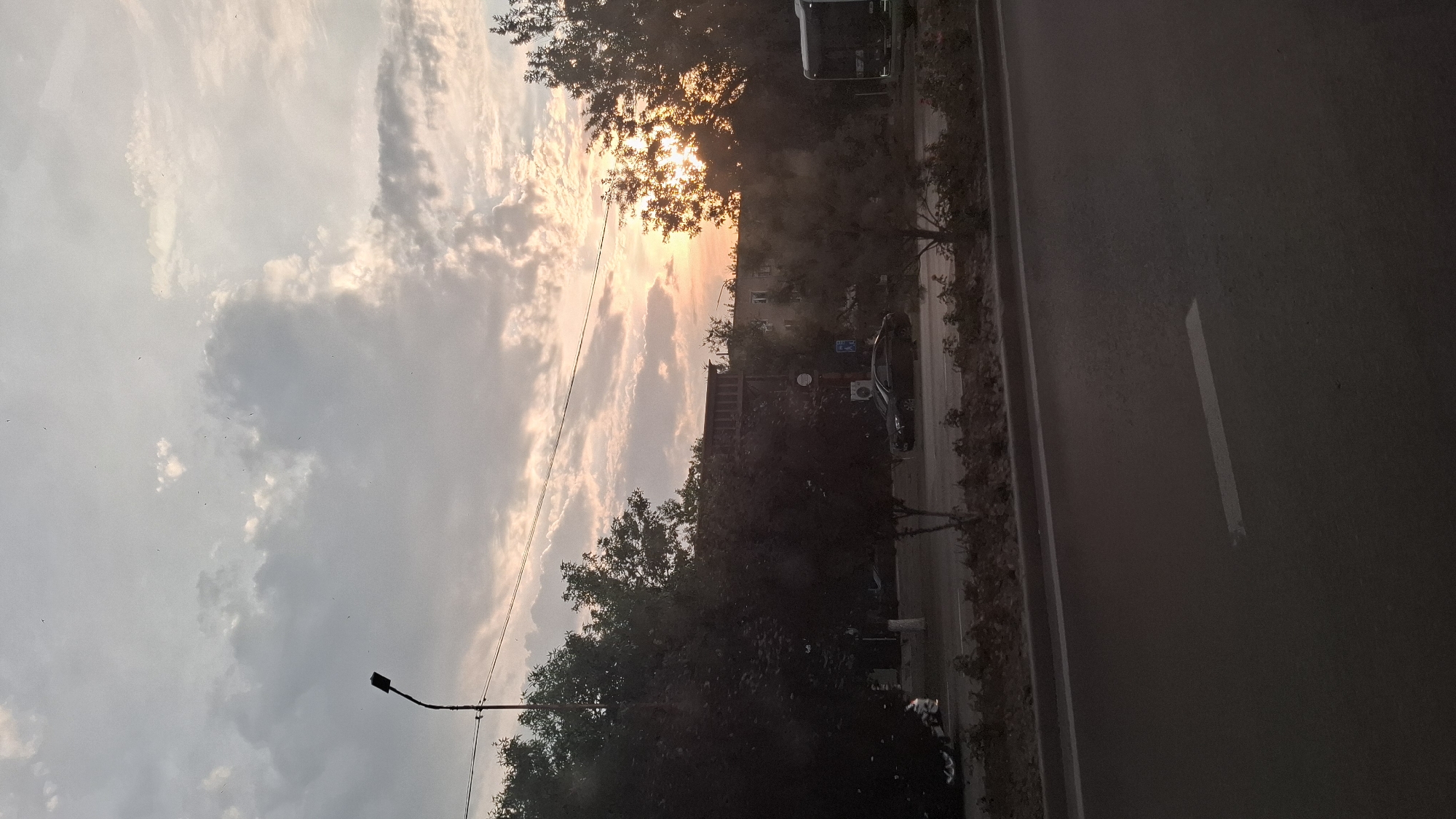 The image depicts a scene captured at sunset in Shymkent, Kazakhstan. The sky is filled with dramatic clouds, tinged with warm hues of orange and pink as the sun, partially obscured by trees, sets. The light creates a soft, diffused glow across the sky.
In the foreground, a street with a white line marking its edge runs vertically, suggesting a road or pavement. To the left of the street, a line of trees creates a dark silhouette against the brightly lit sky, with some buildings and structures visible behind them. A street lamp with a single light fixture is also visible on the left side of the frame.
Further down the street, a dark-colored car is parked, and some scattered debris or trash can be seen along the roadside. The overall impression is that of an urban or suburban setting bathed in the soft, warm light of a fading day. The weather appears to be clear with scattered clouds. There is no visible text in the image.