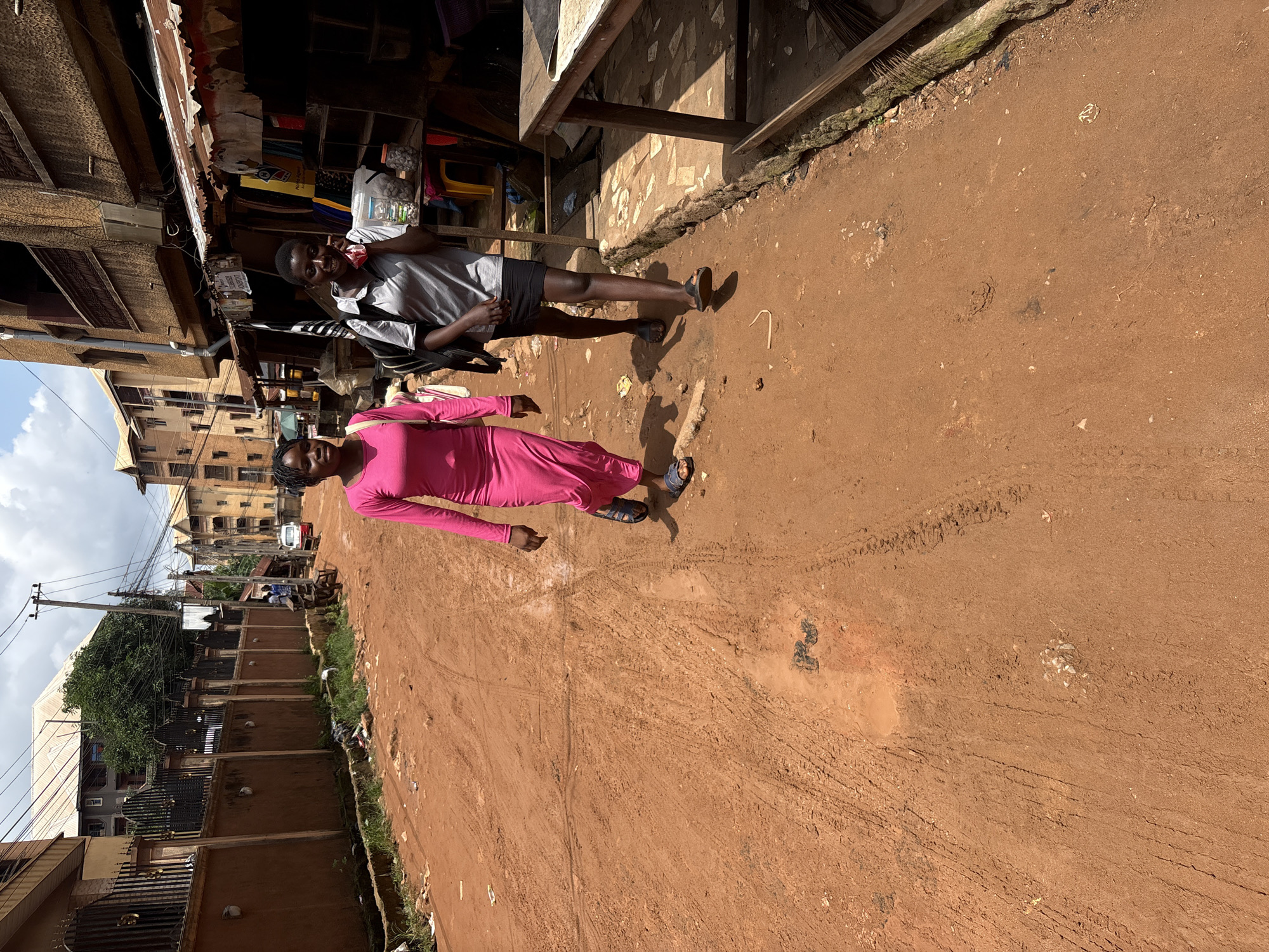 Two young Black women walk down a dusty, reddish-brown dirt road in Nkpor, Nigeria. The woman on the left wears a long-sleeved, bright pink dress and blue sandals. She has her hair pulled back and is looking forward. The woman on the right wears a grey short-sleeved shirt, a black skirt, and black sandals. She has a black backpack on her shoulders and is holding a phone to her right ear, looking towards the camera. They are walking away from the viewer, towards a row of buildings on the right side of the street. These buildings appear to be shops with awnings and open fronts displaying various items. On the left side of the road, there is a brown wall with a black metal fence, and further down the street, there are more residential buildings and a white vehicle. The sky is partly cloudy.