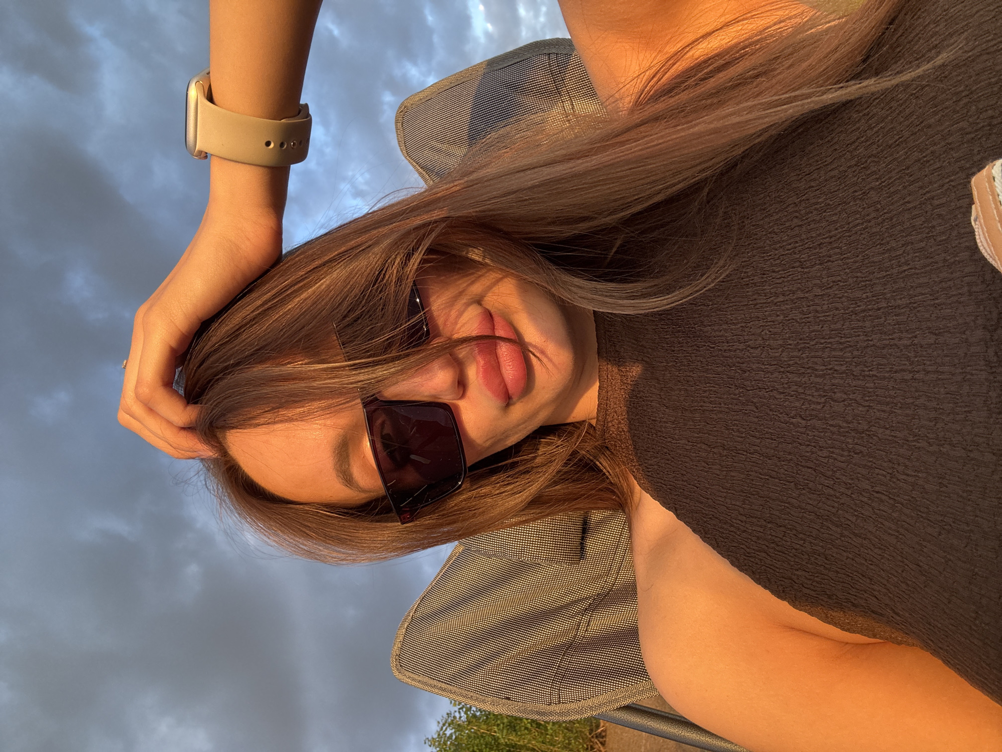 The image is a close-up, low-angle selfie of a young woman taken outdoors during the golden hour, likely late afternoon or early evening, given the warm, directional sunlight illuminating her. She is seated, with a portion of a chair visible in the background. Her left arm is raised, with her hand resting on her head, revealing a light-colored smartwatch on her wrist. She is wearing a dark, textured top. Her hair is long and light brown, framing her face and partially obscuring her eyes, which are covered by dark sunglasses. Her lips are slightly parted, and her expression appears relaxed and possibly playful. The sky in the background is a mix of gray clouds, suggesting a transition in weather, possibly indicating dusk. There is a hint of greenery at the bottom of the frame, suggesting the presence of trees or bushes. The overall mood is casual and summery, with a sense of personal moment captured. No visible text is present in the image. The location context of Nizhny Novgorod, Russia, aligns with the possibility of outdoor activities during pleasant weather.