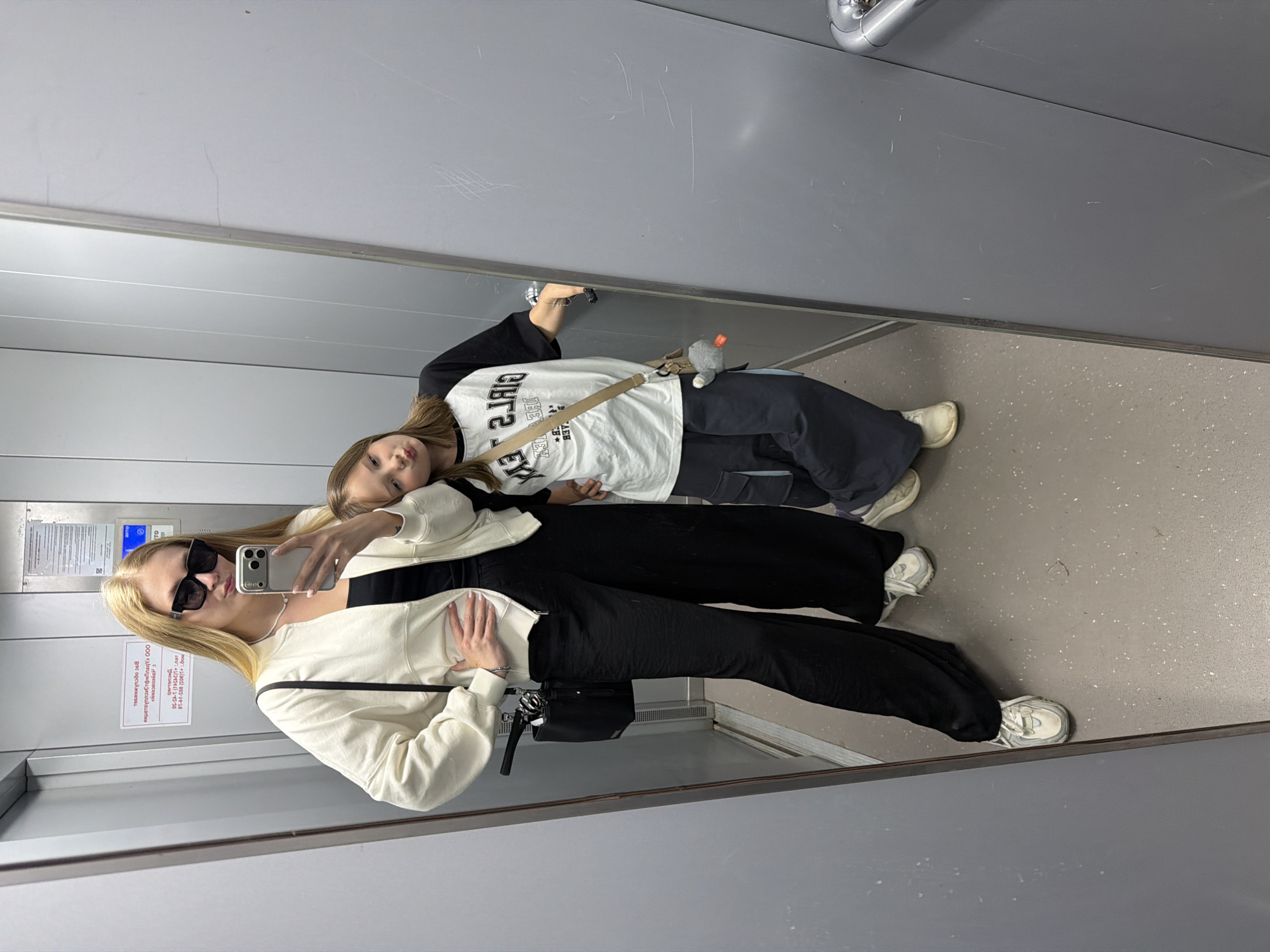 In an elevator, a woman with blonde hair, wearing black sunglasses, a white zip-up jacket, a black top, and black wide-leg pants, poses for a selfie. She has one hand on her hip. Beside her, a younger girl with brown hair, wearing a black and white raglan t-shirt and dark pants, leans forward with her hands on the elevator wall. Both individuals are wearing sneakers. A black purse with a chain strap hangs from the woman's shoulder, and the girl has a tan crossbody bag with a stuffed animal charm. The elevator walls are grey metal, and a notice is affixed to the back wall. The floor is a speckled grey. This scene is in the city of Chaikovsky, Russia.