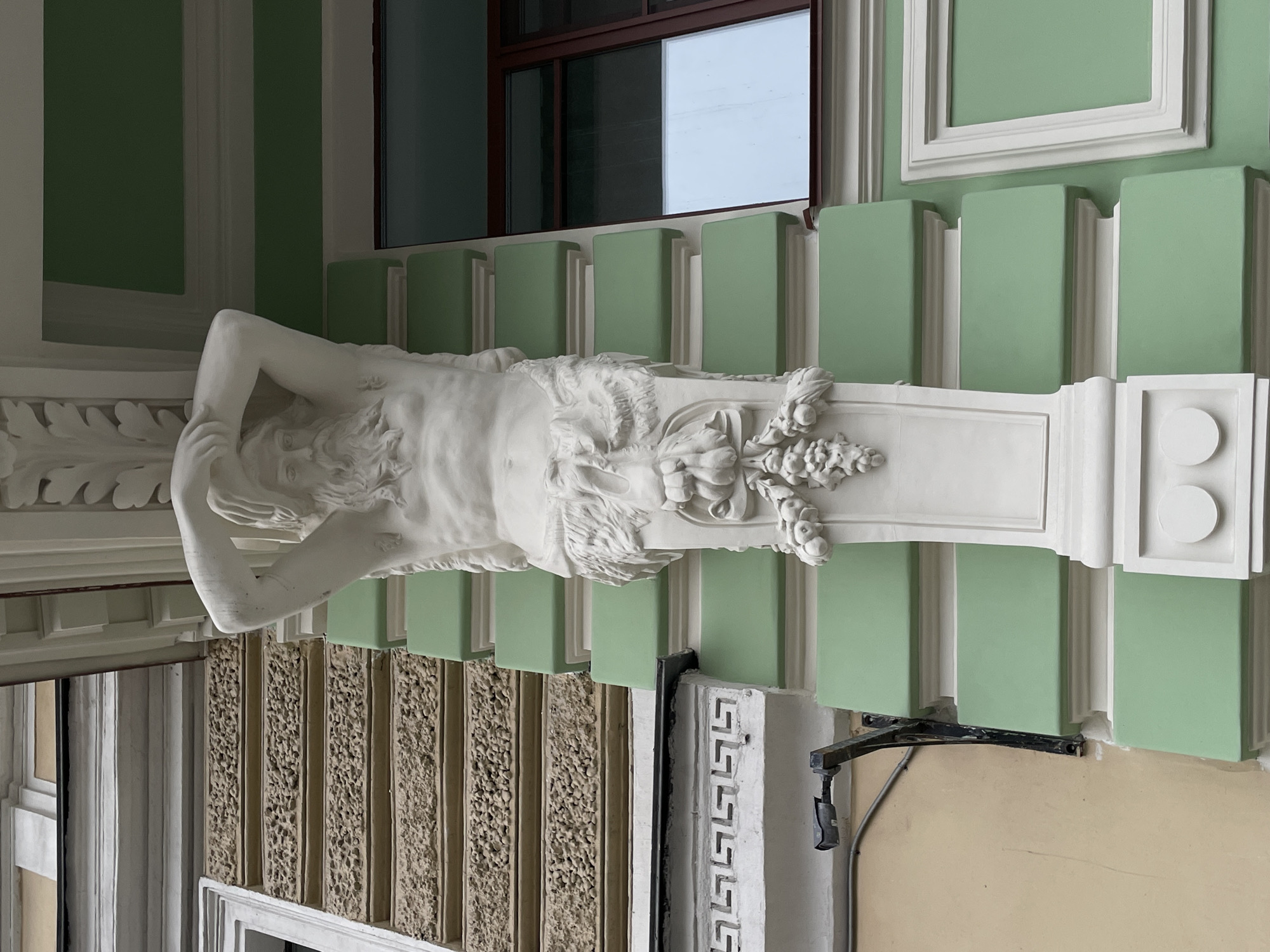 The image shows an architectural detail, specifically a telamon (or atlas) sculpture acting as a supporting element, likely part of a building's facade. The statue is white and appears to be made of marble or plaster. It depicts a male figure, possibly mythological or symbolic, in a strained posture as if bearing a heavy weight. The telamon is mounted on a white pedestal with decorative elements.
The background consists of light green wall segments separated by white vertical stripes. Above the statue, there is a window with a dark brown frame. To the left and below are other architectural details including decorative moldings and a textured wall surface. A black metal bracket is attached to the wall, possibly for a light fixture or sign.
Based on the architectural style and available information, it is highly likely that this image was taken in Saint Petersburg, Russia. The city is known for its neoclassical architecture and elaborate facades, featuring such decorative elements as telamons.