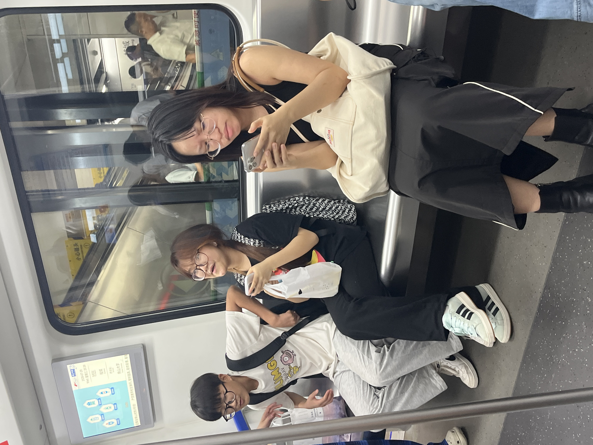 The image depicts three young individuals, two females and one male, seated on a subway train in Chancheng District, China. The male, wearing glasses and a white t-shirt with a graphic print, sits to the left, looking forward and slightly to his right. A young woman in the center, also wearing glasses, holds a white plastic bag in her left hand and a mobile phone in her right, her gaze directed towards the phone. To the right, another young woman, wearing glasses, is engrossed in her mobile phone, which she holds with both hands. She has a large beige tote bag on her lap. All three are seated on a metallic bench. The train's interior features a window reflecting an escalator and other passengers outside, indicating a station environment. A digital display screen mounted on the wall shows Chinese characters and graphics related to public transportation information, including dates and times. The lighting suggests an indoor, possibly underground, setting.