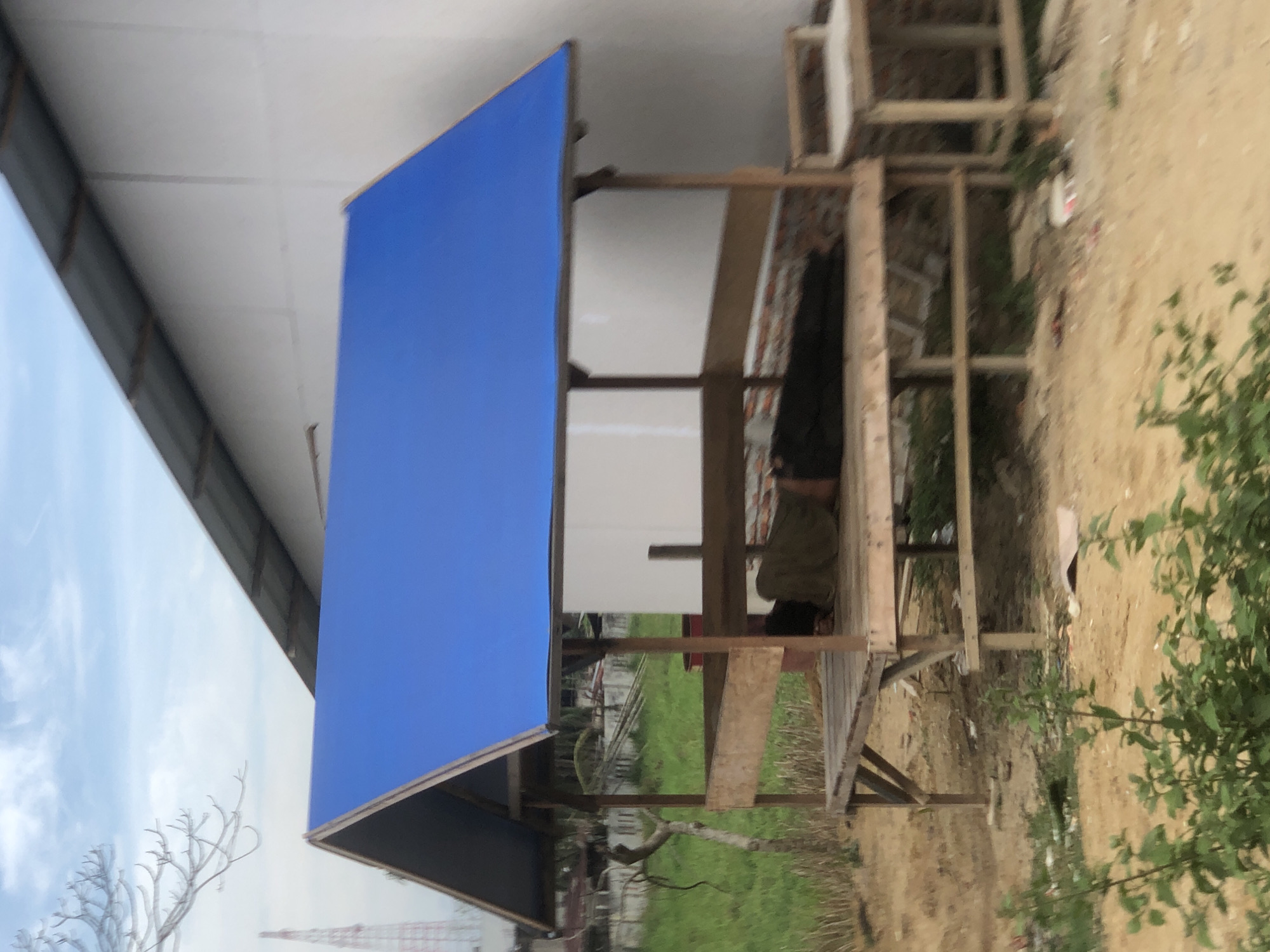 A medium shot captures a partially constructed wooden structure with a vibrant blue awning. The structure is situated near a building with a white exterior wall, suggesting an urban environment. A person is present within the wooden frame of the structure. The surrounding area consists of vegetation and bare ground, indicative of a site undergoing construction or development. The location is Teupinbatee, Indonesia.