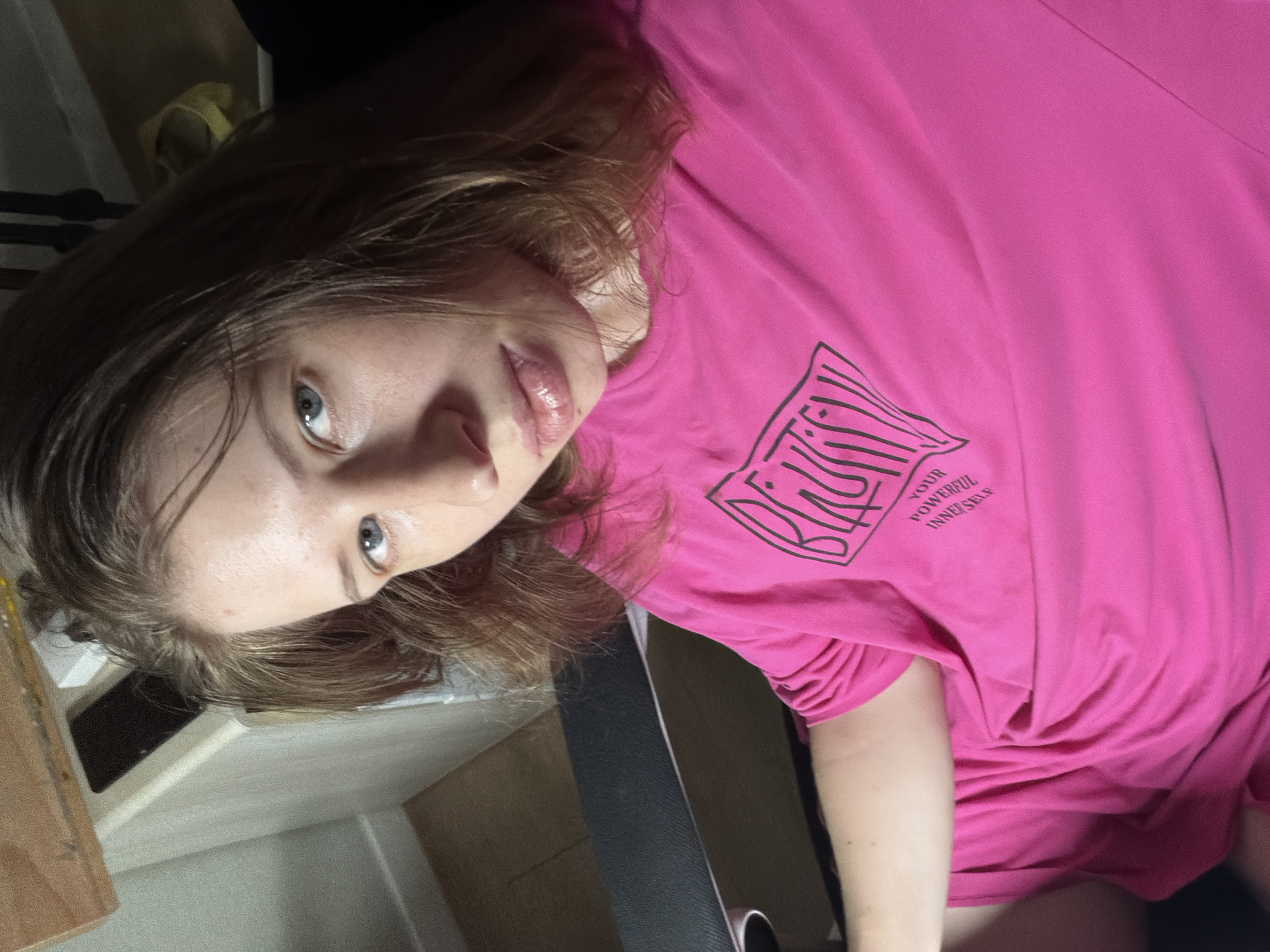 A young woman with light brown hair and blue eyes is seated, looking directly at the camera. She is wearing a bright pink t-shirt with a black graphic design and text that reads "YOUR POWERFUL INNER SELF". The background suggests an indoor setting, possibly a room with furniture visible in the upper left corner. There is no indication of the city of Dongducheon-si, South Korea.
