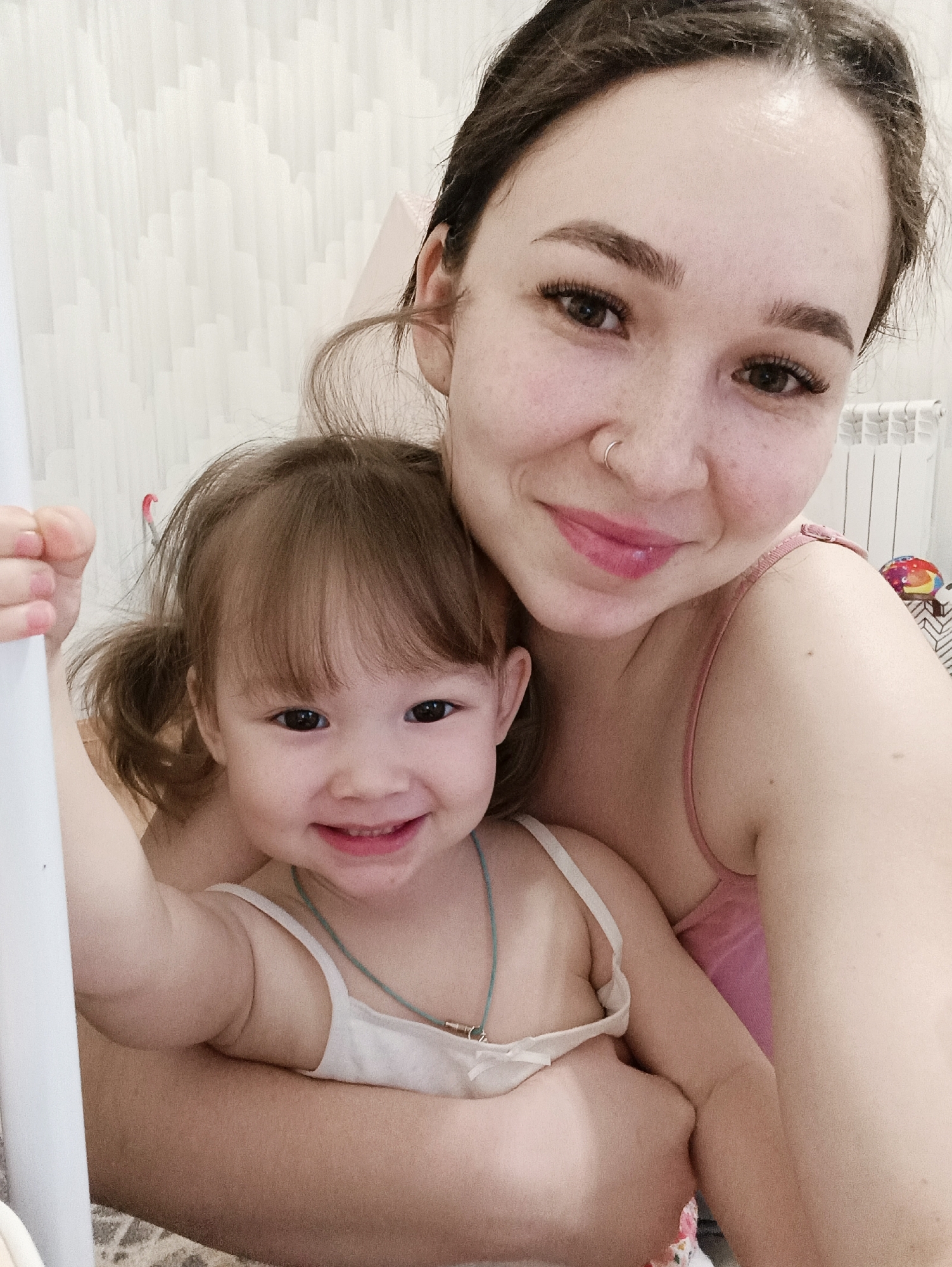 This image, captured in Izhevsk, Russia, displays an adult female and a young female child in an indoor setting. The adult, positioned to the right, has dark hair, dark eyes with prominent eyelashes, a nose ring in her right nostril, and wears a pink strap top. She exhibits a closed-mouth smile. The child, a toddler, is positioned in front of the adult, with light brown hair styled in pigtails and bangs, dark eyes, and a broad smile. She wears a white strap top and a light blue cord necklace with a small metallic pendant. Her left hand is observed gripping a white vertical bar, possibly part of a bed or playpen structure. The background consists of a light-colored wall with a subtle, geometric texture. A white radiator is visible on the far right, and a colorful, indistinct object is partially visible above it. A patterned textile is partially visible along the bottom edge of the frame.