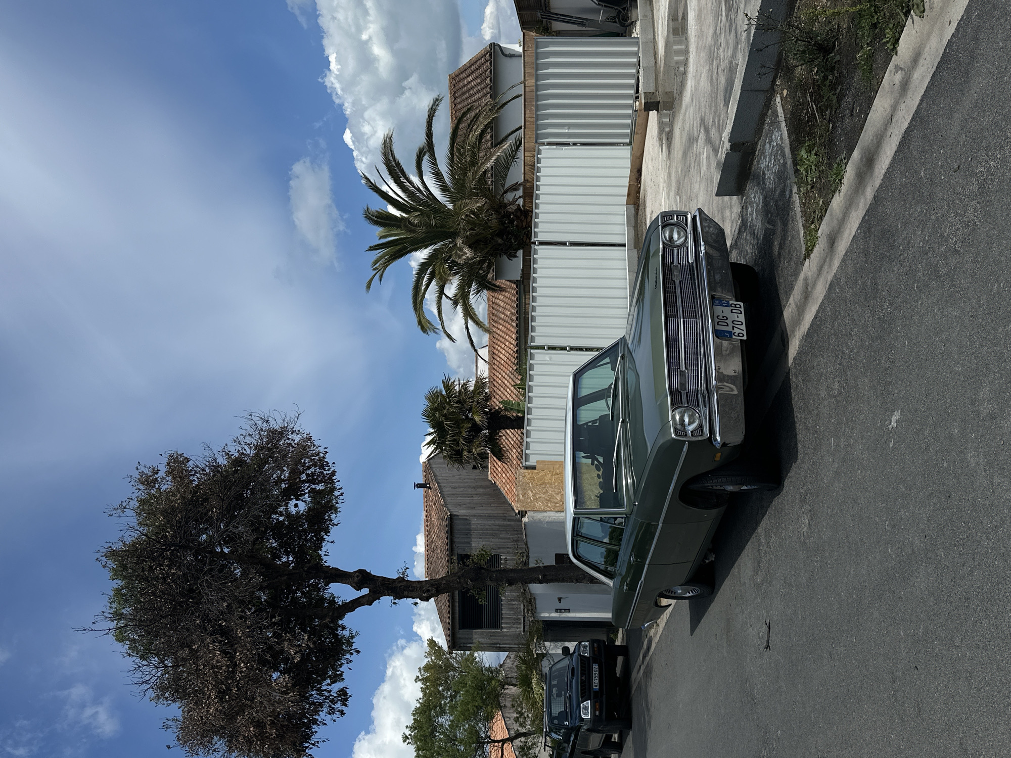 The image shows a residential street scene in La Couarde-sur-Mer, France. A vintage green car is parked on the right side of the road, facing away from the viewer. The car's license plate, "DG-670-DB," is visible. To the left of the car is a white fence, behind which are buildings with tiled roofs and a palm tree. Further down the street is a dark-colored SUV. A large tree with sparse foliage is located on the left side of the street, under a blue sky with scattered clouds. The road surface is asphalt.