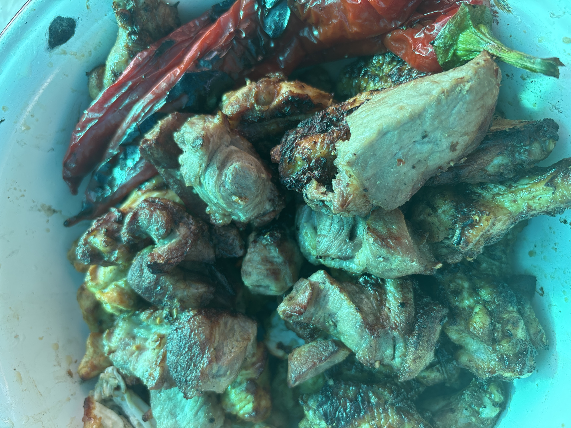 The image shows a close-up, overhead view of a white container filled with cooked meat and red bell peppers. The meat appears to be grilled or roasted, with some pieces showing charring and crispy edges. The peppers are whole and appear to be roasted as well, with some browning and wrinkling. The lighting in the image is bright and casts a turquoise hue over the food. There are no people or any discernible environmental details that would indicate a specific location in Zheleznogorsk, Russia. The scene is a still-life of food, with no actions or interactions taking place.
