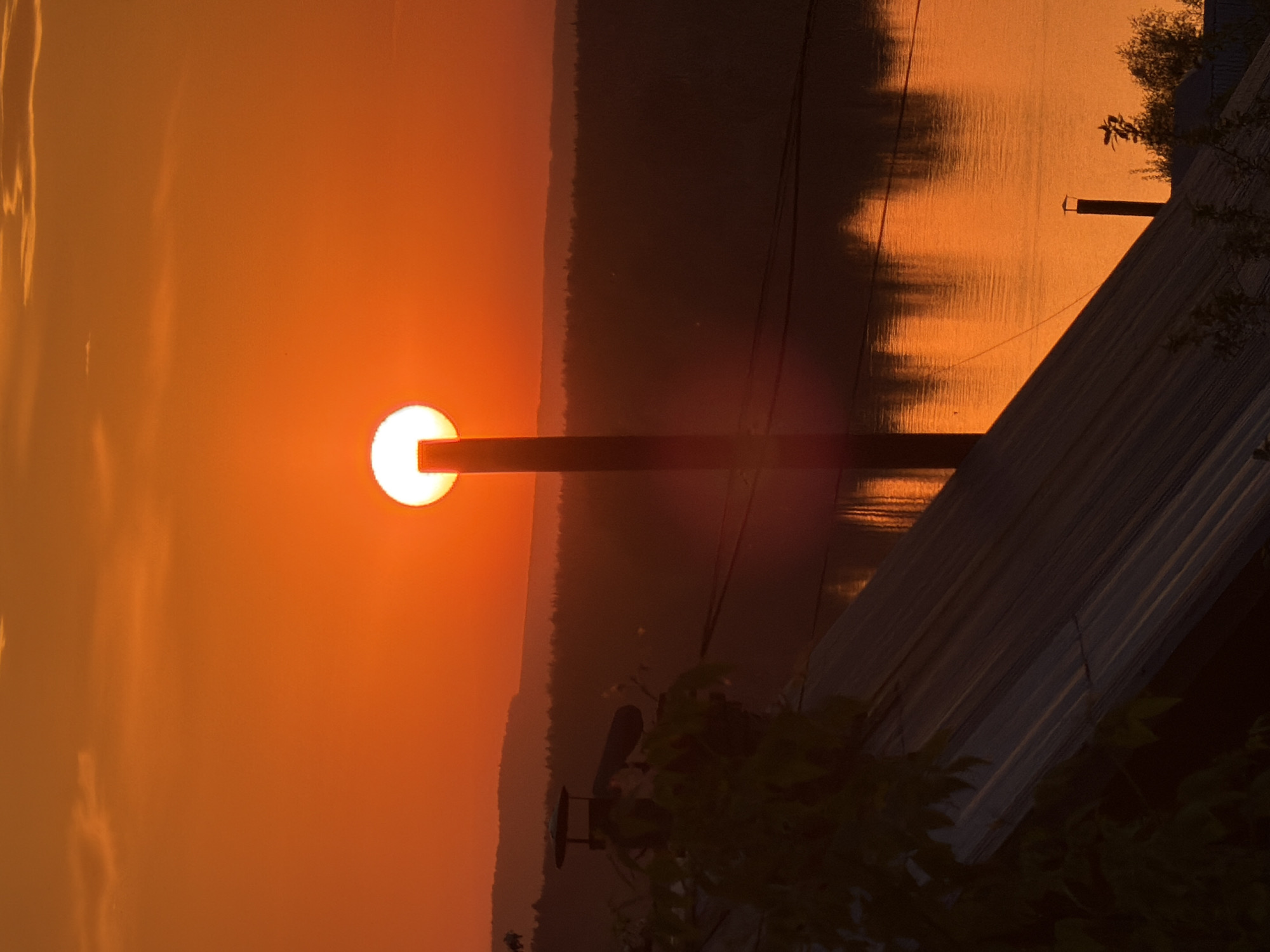 The image depicts a sunset in Chaikovsky, Russia. The sun, a bright orange orb, is partially obscured by a vertical pole, possibly a power line support. A body of water, likely the Kama River, reflects the orange hues of the sunset. In the foreground, a corrugated metal roof and foliage are visible, suggesting the image was taken from a building or elevated position. The sky is a gradient of orange and yellow, fading into darker tones near the horizon. Trees and vegetation line the distant shore. Power lines are visible stretching across the river.