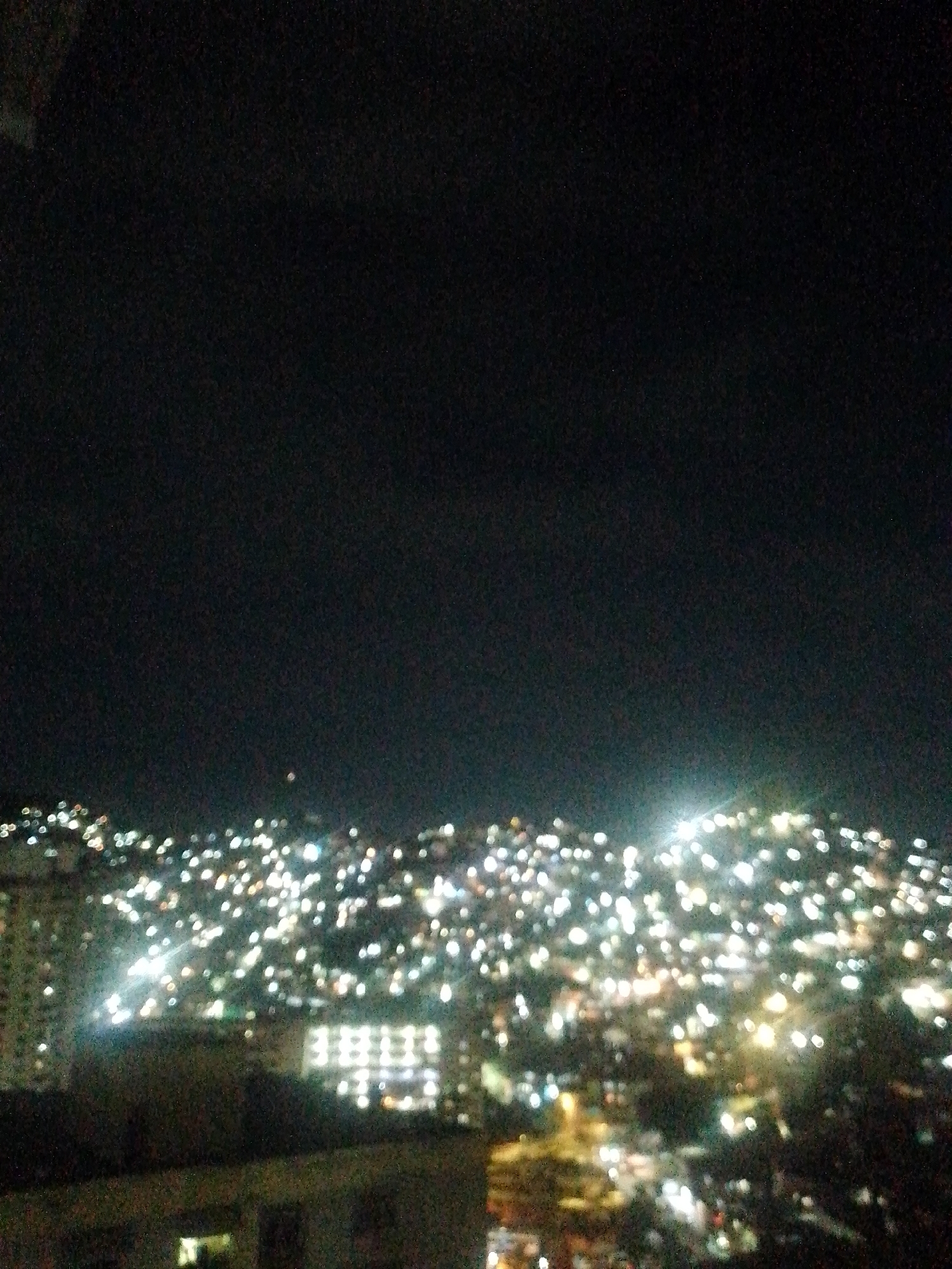 The image shows a nighttime view of a hillside city, likely Caracas, Venezuela, illuminated by numerous lights. The scene is somewhat blurry, with the lights appearing as bokeh. In the foreground are buildings with illuminated windows. The rest of the image is dominated by the dark sky and the dense cluster of lights extending across the hillside. No people or specific actions are visible.
