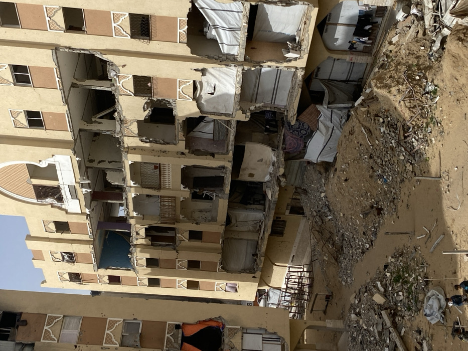 This image depicts a devastated urban residential area in Khan Yunis, Palestinian Territory, under clear daylight conditions. Two multi-story buildings, constructed with light brown exteriors and decorative elements, show extensive structural damage. The central building, appearing to be an apartment block, has multiple floors with outer walls and interior partitions severely compromised or completely missing, revealing exposed concrete slabs and twisted rebar. Many openings are covered with makeshift white or patterned tarps and sheets. Clothes are seen hanging from balconies and within damaged sections of the lower floors. The exterior walls of both buildings exhibit numerous small impact marks, consistent with bullet holes. The ground between and in front of the buildings is covered with substantial piles of sand, concrete rubble, and miscellaneous debris. In the bottom left foreground, four individuals, appearing to be adult males or older children, are visible on the debris-strewn ground. Their specific activity is not discernible, but they are situated amidst the destruction.
