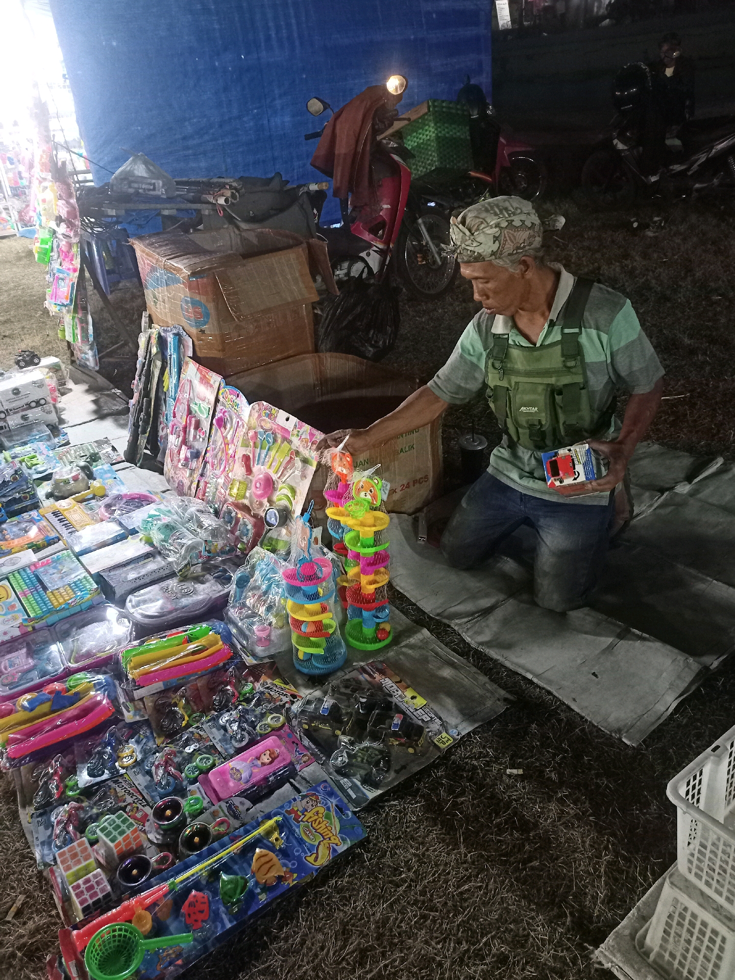 The image depicts an outdoor stall in Tanon, Indonesia, likely at night. A man, wearing a light-colored striped shirt, a green vest with multiple pockets, and a patterned bandana, kneels on a grey tarp. He holds a small, red and white toy box in his right hand. The stall is filled with various children's toys, including toy fishing rods, toy cars, Rubik's cubes, and colorful stacking toys. Some toys are packaged in plastic, while others are loose. Behind the stall, cardboard boxes are visible, along with a motorcycle with a red seat and a green basket. A large blue tarp is stretched out in the background, providing shelter. The ground is covered with grass and dry leaves. Several other motorcycles are parked nearby. A white plastic basket is stacked on the right side of the image. The scene suggests a lively night market or a temporary outdoor sales event.