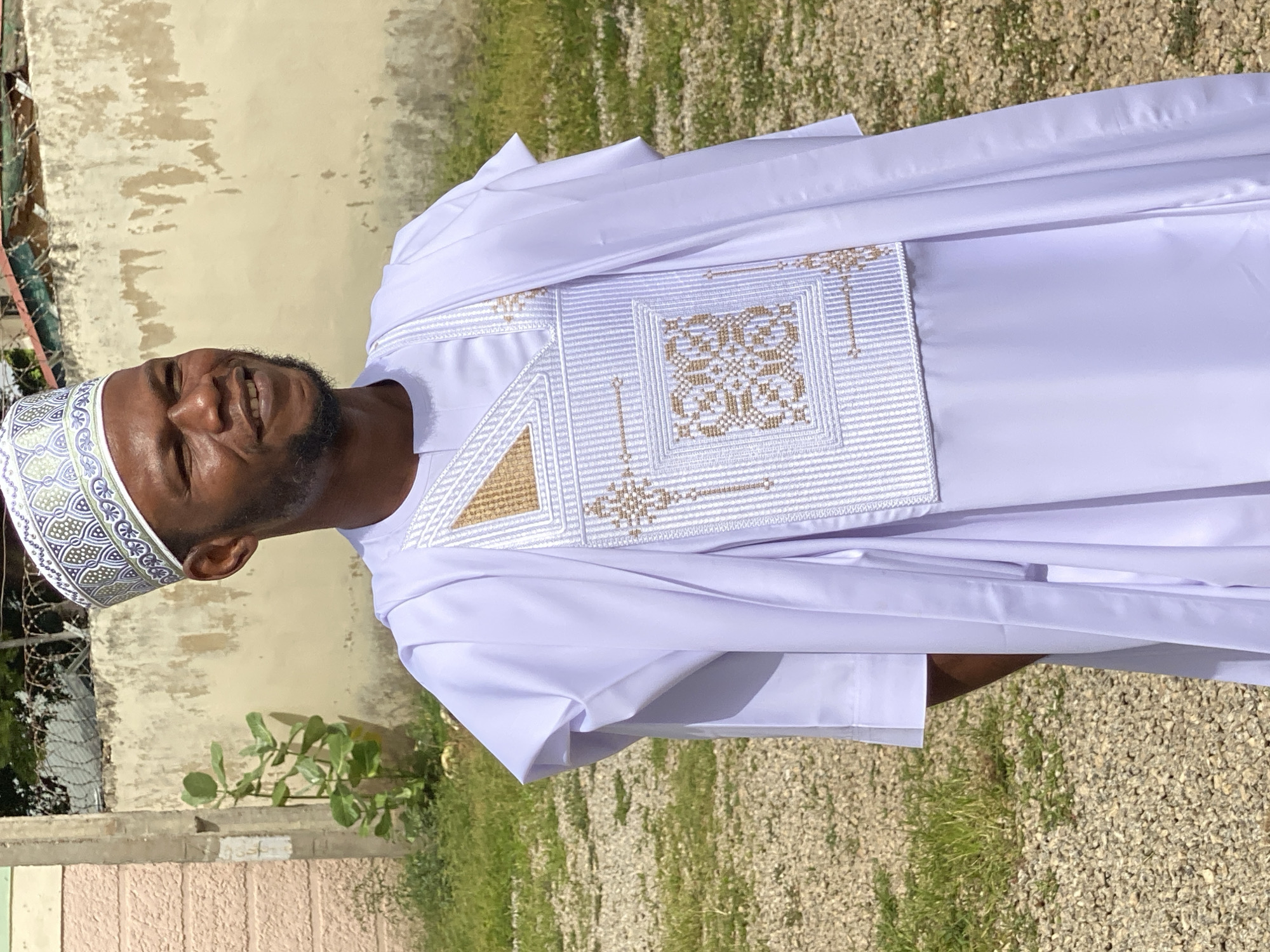 The image shows a man wearing a traditional white thobe and a patterned Kufi hat. He is smiling broadly, with his eyes closed, suggesting happiness or amusement. The thobe is adorned with intricate white and gold embroidery on the chest. The man has a beard and is looking towards the viewer.
The setting appears to be outdoors in a sunny environment, as indicated by the bright lighting and the shadows. There is a wall with a weathered texture behind him, and an area with green grass and gravel in the foreground. The overall impression is one of a pleasant day, possibly in a warm climate. The man's attire suggests a cultural or religious context, perhaps for a celebration or a religious gathering. The image is taken at an angle, with the subjects appearing sideways. There is no visible text in the image. The location context provided is Dadi Riba, Nigeria.