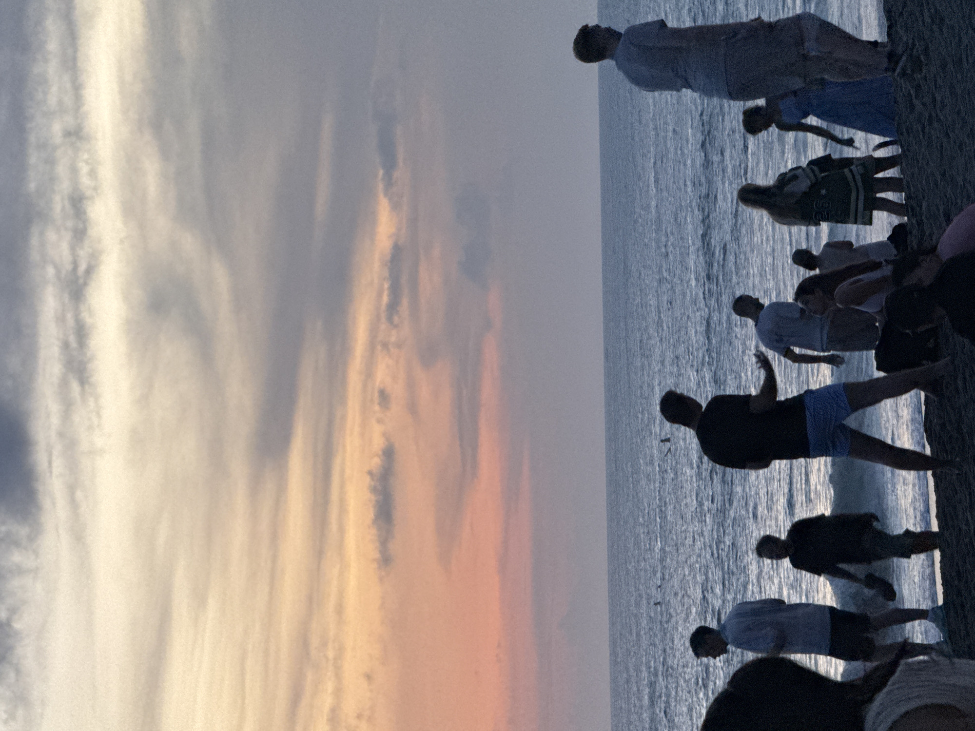 The image depicts a group of people standing on a beach at sunset. The location is likely North Kuta, Indonesia, given the lighting and coastal environment. Several individuals are silhouetted against the ocean and sky. The sky features a gradient of colors ranging from gray to orange. The ocean is visible in the right side of the picture. Some people appear to be interacting with each other. A tree is positioned on the right corner of the image.