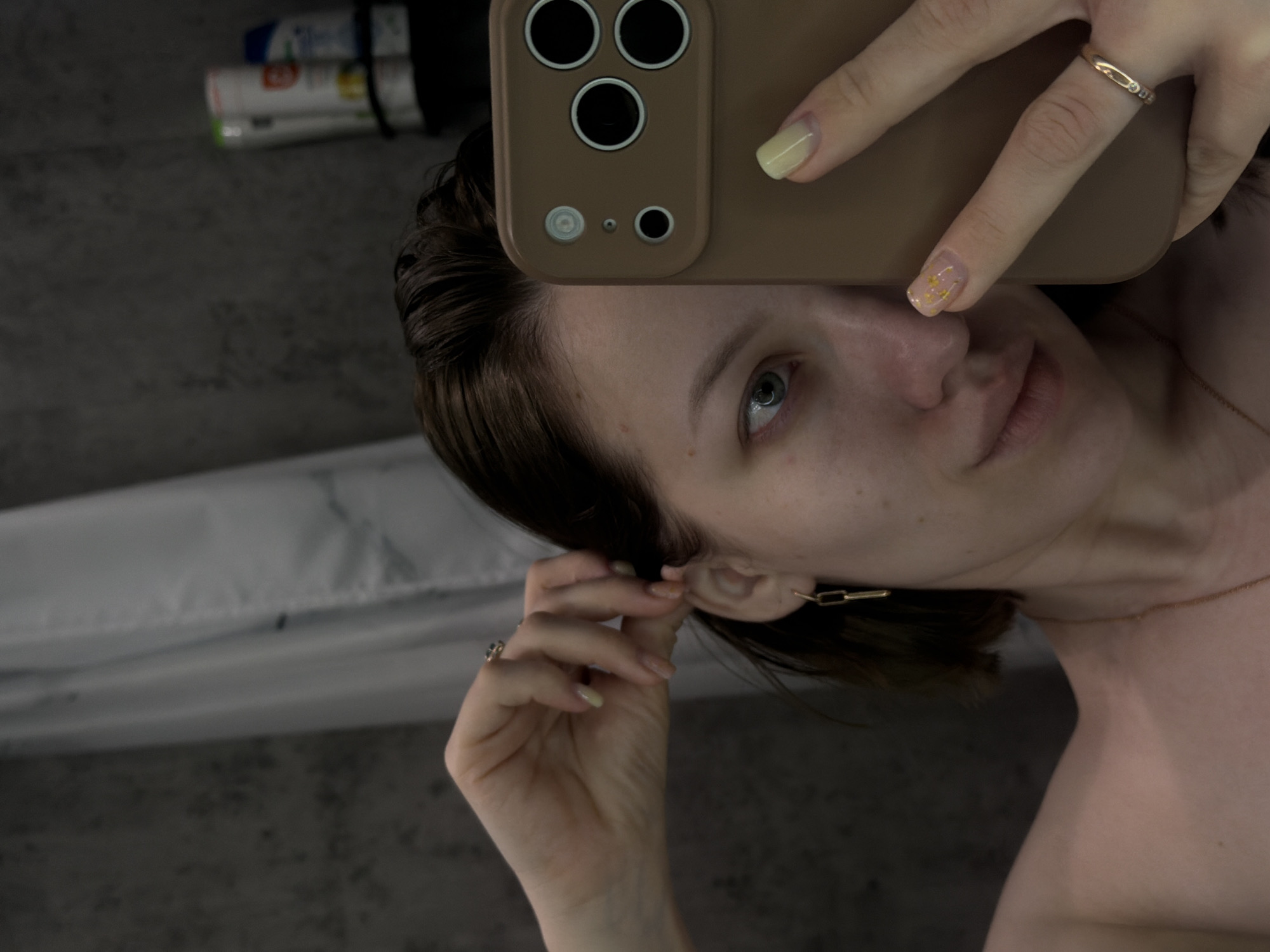 The image shows a person holding a smartphone up to their face, presumably taking a selfie. The phone is a light brown color with three visible camera lenses and a flash. The person has fair skin, dark hair, and blue eyes. They are wearing gold jewelry, including a ring on one finger, earrings, and a necklace. The background is a neutral gray color, possibly a wall, with a white object (possibly a towel or fabric) visible in the lower-left corner. There are also toiletry bottles on the top left. There is no specific information in the image to indicate the location is Reshetnikovo, Russia.