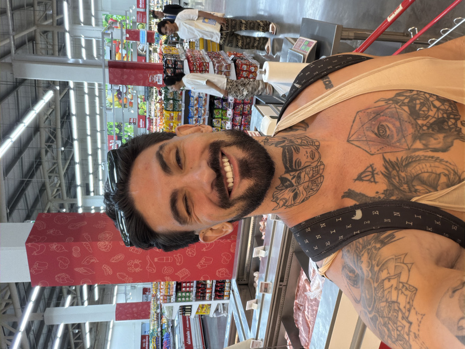 A male individual with dark hair, a beard, and numerous tattoos on his neck, chest, and left arm, is depicted from the chest up, smiling broadly and winking with his left eye. He wears a light-colored tank top and a dark backpack strap is visible over his left shoulder. Black sunglasses rest on top of his head.
The scene is a large supermarket, specifically a fresh food section in Thailand, identifiable by Thai script on overhead signage and product displays. Immediately behind the man is a refrigerated display case containing raw meat, including cuts resembling pork belly. To his right, on a stainless steel counter, are a roll of paper towels and a digital weighing scale. In the background, multiple aisles are visible with shelves stocked with various packaged goods, and further back, a brightly lit produce section. A prominent red pillar adorned with white outline graphics of food items (vegetables, meats, beverages) stands in the center-left. Overhead, the ceiling features industrial-style fluorescent lighting.
Three other individuals are present in the background: one person in a white top and patterned trousers on the left, another male in a white top and patterned trousers carrying a white shoulder bag on the right, and a partially visible person in a dark suit jacket further to the far right. The flooring is grey tile. This establishment is consistent with a major hypermarket chain found in the city of Bangkok, Thailand.