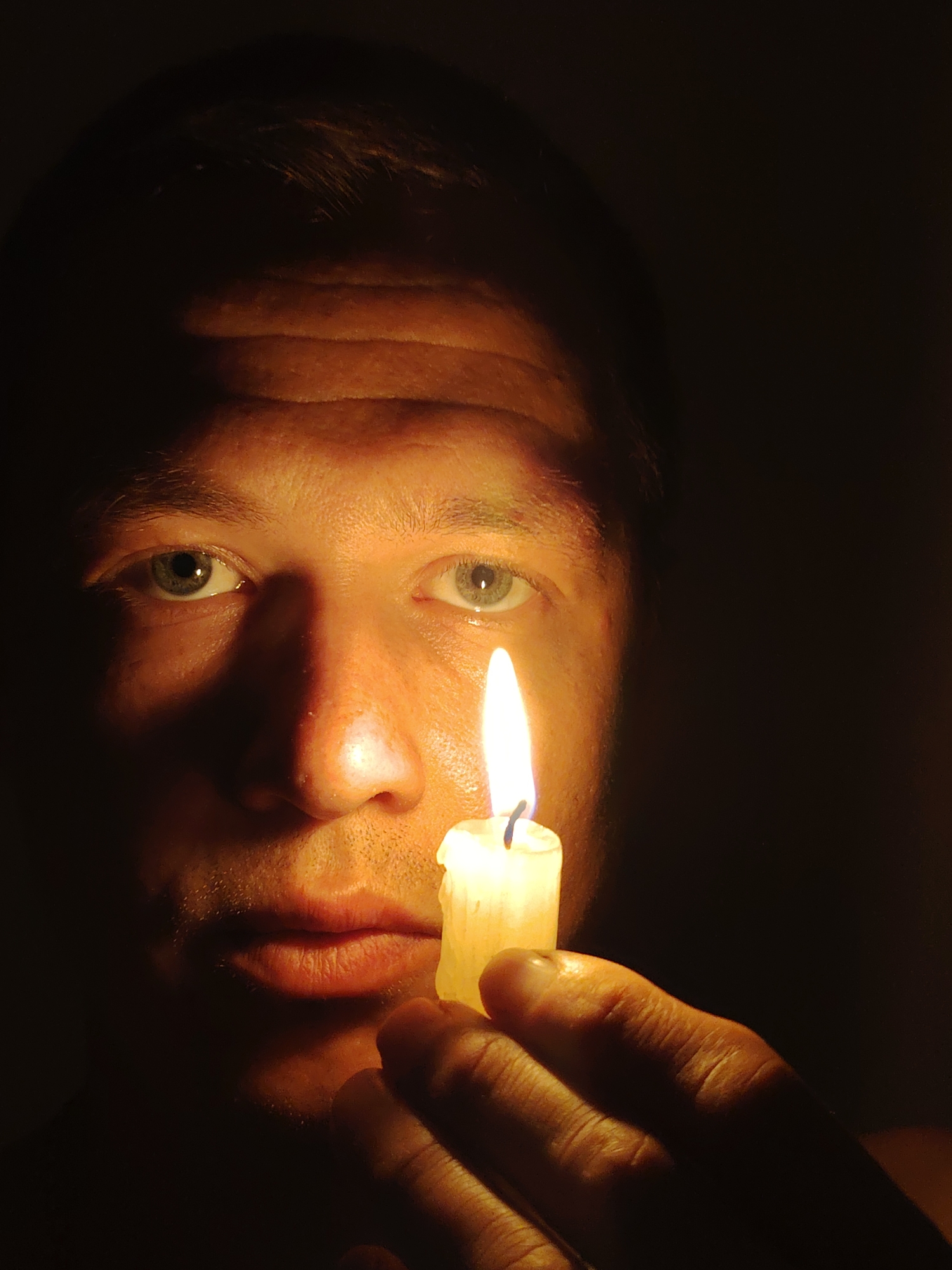 A close-up, waist-up shot captures a man holding a lit candle in the foreground, his face illuminated by its warm glow against a dark background. The man, positioned to the left of the frame, is looking towards the candle with his eyes reflecting the flame. His skin is tanned, and his short hair is visible. The candle, held in his right hand, is positioned near his chin, casting a strong light on his features and creating a dramatic shadow across the left side of his face and forehead. The right side of his face is fully illuminated by the candle. His mouth is slightly open, and his lips are moist. The background is uniformly dark, offering no discernible environmental details or location. There is no information to suggest this image is located in Chaikovsky, Russia.