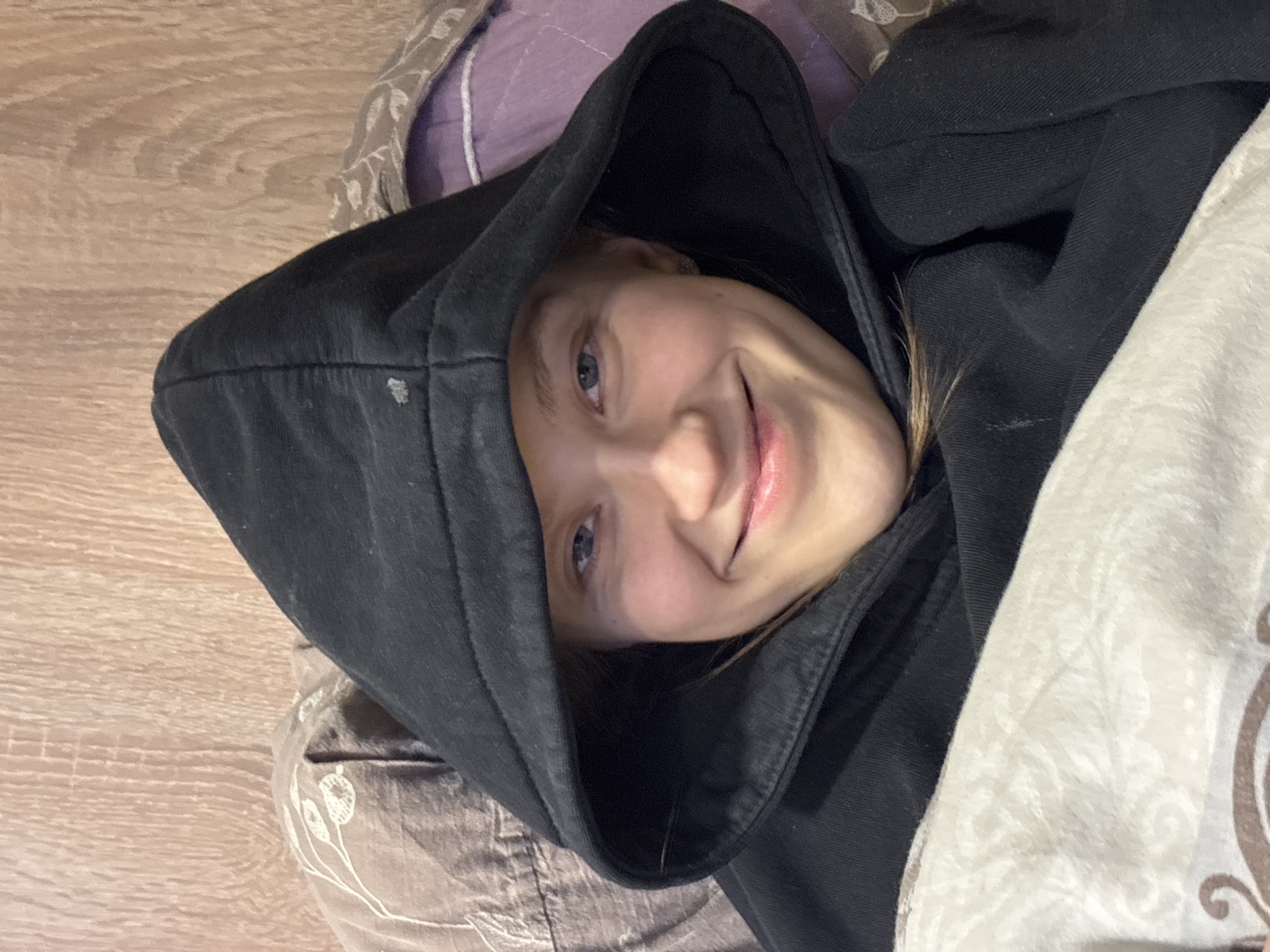 A young person wearing a black hooded sweatshirt lies on what appears to be a bed or similar surface. The person is smiling at the camera. Parts of a pillow or bedding are visible in the background, including colors of purple, beige, and brown. The surface below the person has a wood-grain pattern. There is no evidence in the image that suggests the location of the city of Perm, Russia.