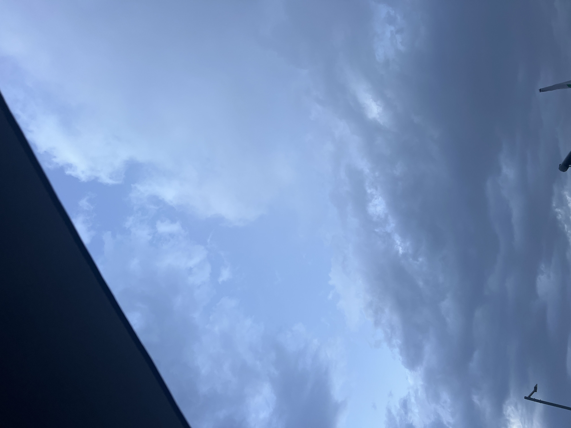 The image shows a cloudy sky with dark clouds gathering. The sky is a mix of blue and gray hues. In the lower left corner, a streetlight is visible. In the lower right corner, a part of an aircraft wing can be seen. The scene appears to be an outdoor environment, possibly an airport or a location near an airport.