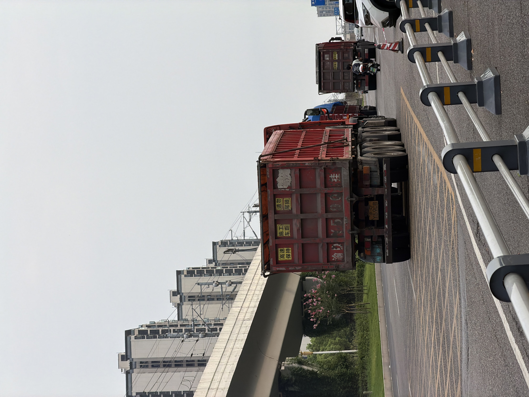 In Jiangjin, China, the image captures a cloudy day on a multi-lane road, likely an urban or suburban highway. A prominent feature is a raised concrete structure, possibly an overpass or elevated railway, running diagonally across the upper left portion of the frame. Along this structure, and in the background, are several modern, multi-story residential buildings, their facades light-colored and punctuated by dark windows.
The foreground is dominated by a red dump truck in the center, facing away from the camera. It has a license plate that reads "Y590╙". Yellow signs are affixed to the back of the truck, one with Chinese characters "高速", "保持", and another with "施工", "车距". To the right of this truck, another, similar dump truck is visible, also facing away, with two people on a motorcycle behind it. Further down the road, a white SUV is partially visible.
The road surface is asphalt, marked with yellow grid lines in the immediate foreground, indicating a no-parking or special zone. Metal barriers with grey bases and light grey railings, accented with yellow rectangles, line the right side of the road. Greenery, including trees with pink blossoms, is visible on the left side of the road, growing beneath the elevated structure. Power lines and poles are also present, running along the elevated structure and behind the buildings. The overall impression is a busy road scene in a developed urban area.