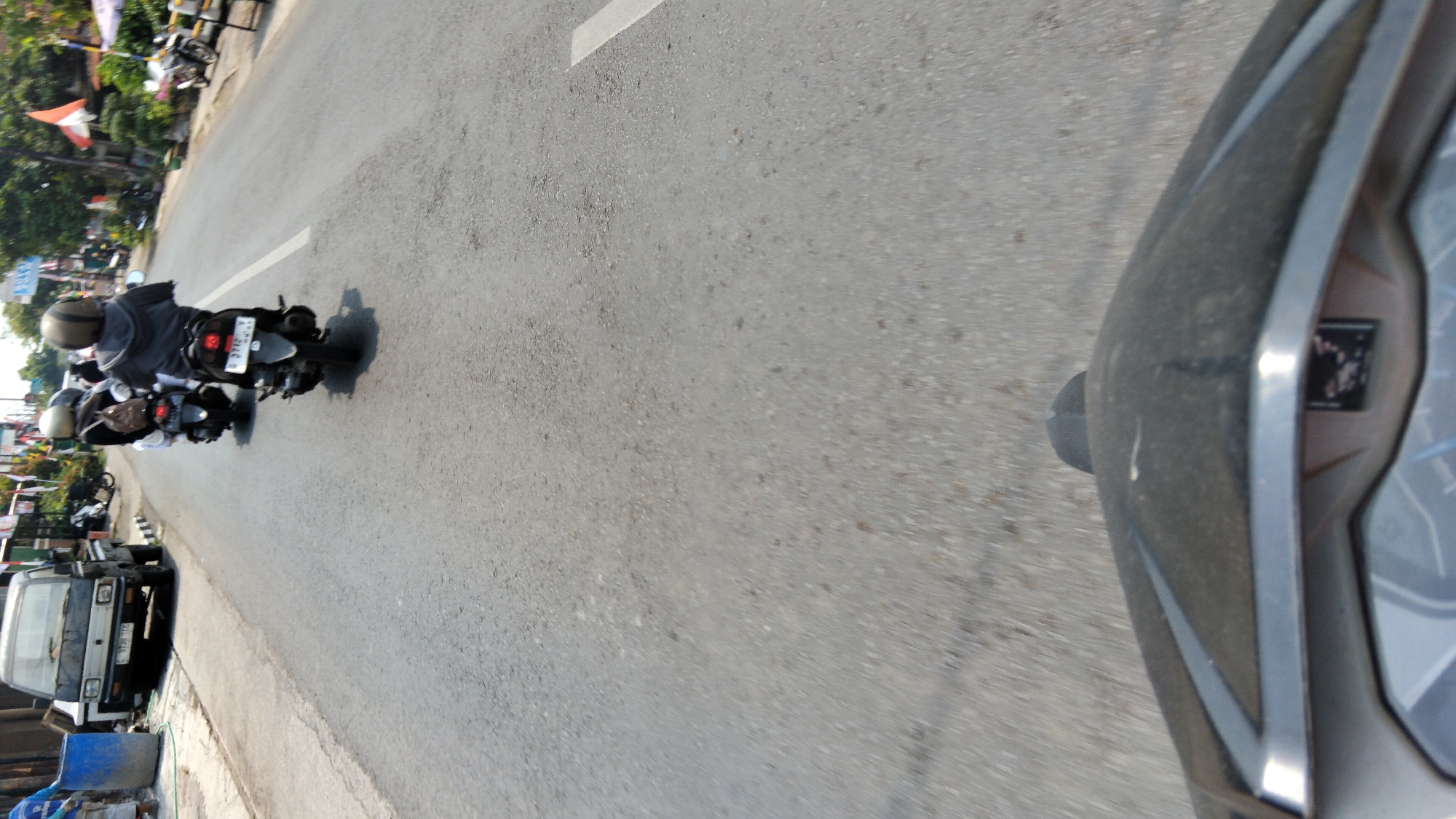 This image depicts a scene from a moving vehicle, likely a motorcycle given the perspective of the handlebars and dashboard in the foreground. The perspective suggests the photographer is traveling down a street in Proyonangg, Indonesia.
In the middle ground, several motorcycles are visible. The closest motorcycle has a rider wearing a gray hoodie and a helmet. Another motorcycle further ahead has a rider in a white shirt and a helmet. Behind them, another motorcycle with a rider wearing a dark jacket and helmet is also visible.
To the left of the road, a small truck is parked. It appears to be a utility vehicle. A blue barrel is also present near the truck. The road itself is asphalt, with a white line marking the lane. The environment suggests an urban or semi-urban setting with buildings and vegetation visible in the background, indicating a typical street scene in Indonesia. The lighting indicates it is daytime.