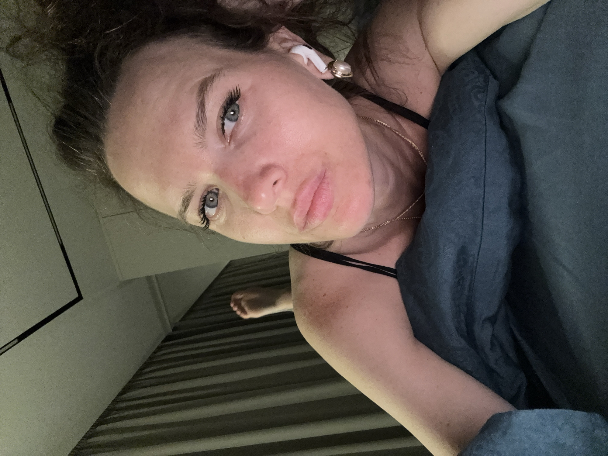 A woman lies in bed with a dark blue blanket pulled up to her chest, wearing a black tank top and a gold necklace. She has curly brown hair, fair skin, blue eyes, and is wearing a white earpiece. The room features striped curtains or blinds, a white wall, and a glimpse of a ceiling corner with a black frame. A single foot is visible on the far side of the bed. There is no indication the location is in the city of Khimki, Russia.