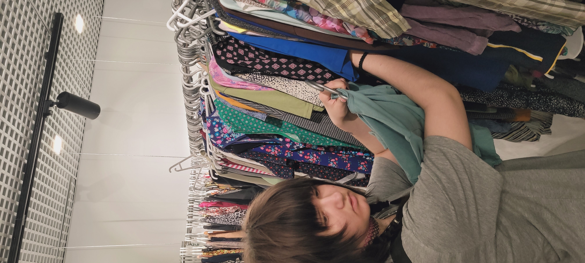 A young person with dark, medium-length hair and bangs, wearing a grey t-shirt and a dark choker necklace, stands in front of a clothing rack. They are focused on examining garments, holding a light teal/green piece of clothing on a silver metal hanger with both hands. The clothing rack is densely packed with a diverse assortment of multi-colored and patterned clothes on various white, grey, and silver hangers. In the background, a white wall is visible beneath a grid-patterned ceiling featuring a black track lighting system with a cylindrical spotlight and recessed square lights. The scene depicts an individual browsing clothes in what appears to be a retail environment in Unknown, Unknown.