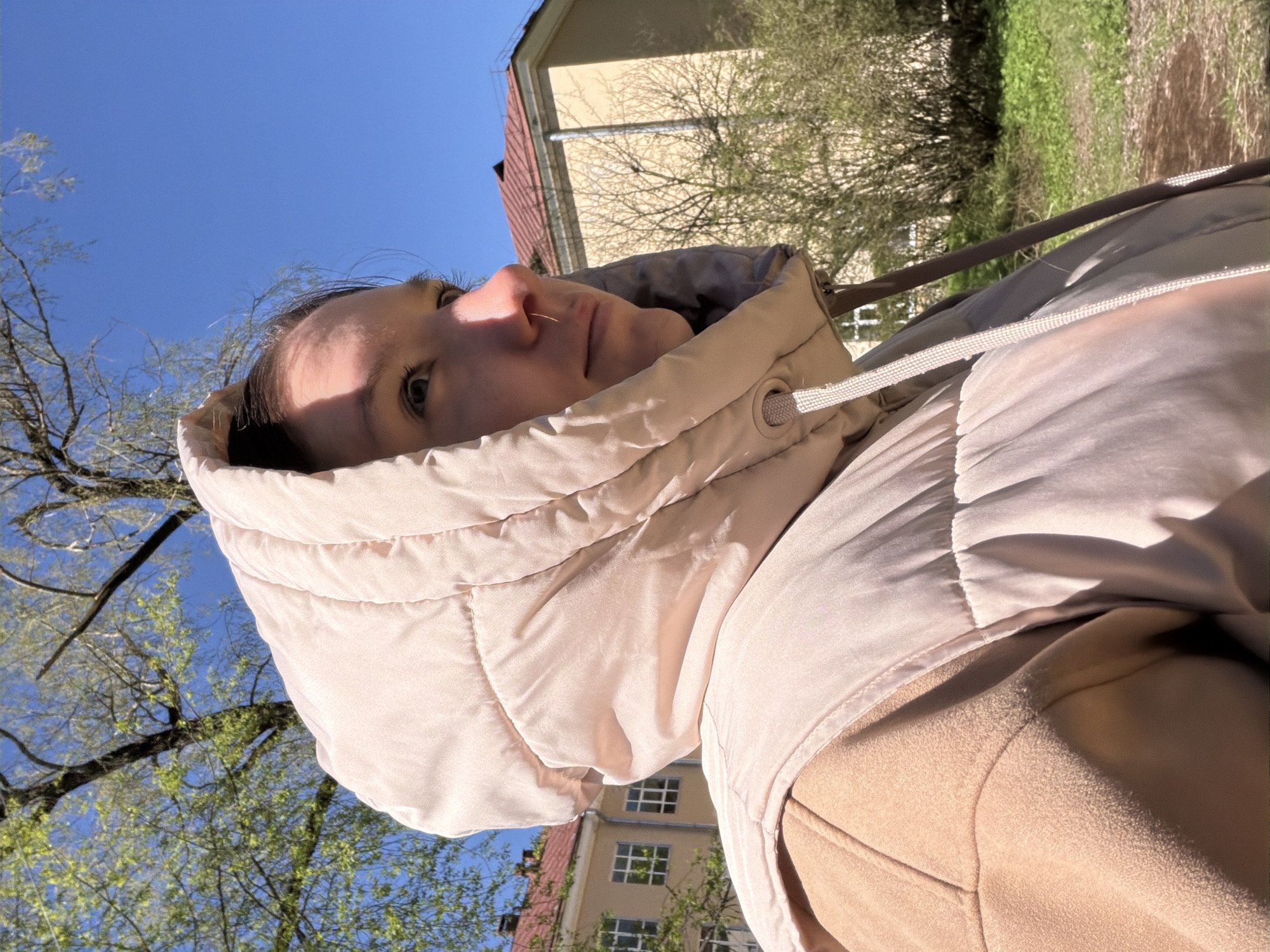 A woman wearing a light beige hooded puffer jacket is outdoors in Tokkari, Russia. Her head is tilted back, and the sky and some trees are visible above her. Buildings are in the background. The lighting is bright, suggesting it is daytime.