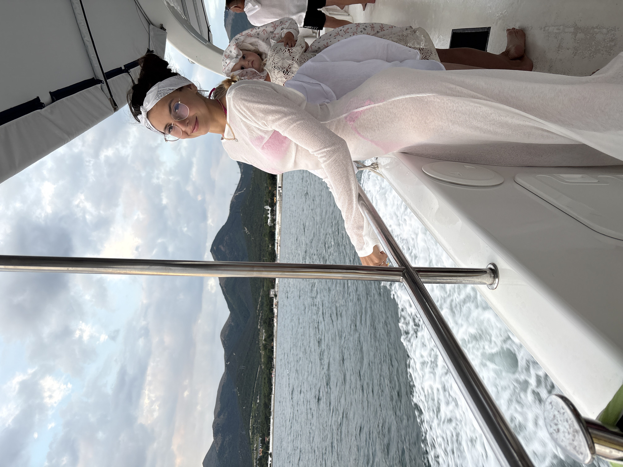 A woman with dark hair tied back with a white bandana and wearing glasses and a sheer white dress over a pink bikini smiles at the camera while standing on the deck of a boat. She is leaning on a metal railing. Behind her, a child in a white lace bonnet and dress is being held by an unseen person. In the background, there are calm waters, mountains, and a cloudy sky. Another person in a white hoodie and dark shorts is visible on the right side of the frame. The boat is moving, as evidenced by the wake behind it.