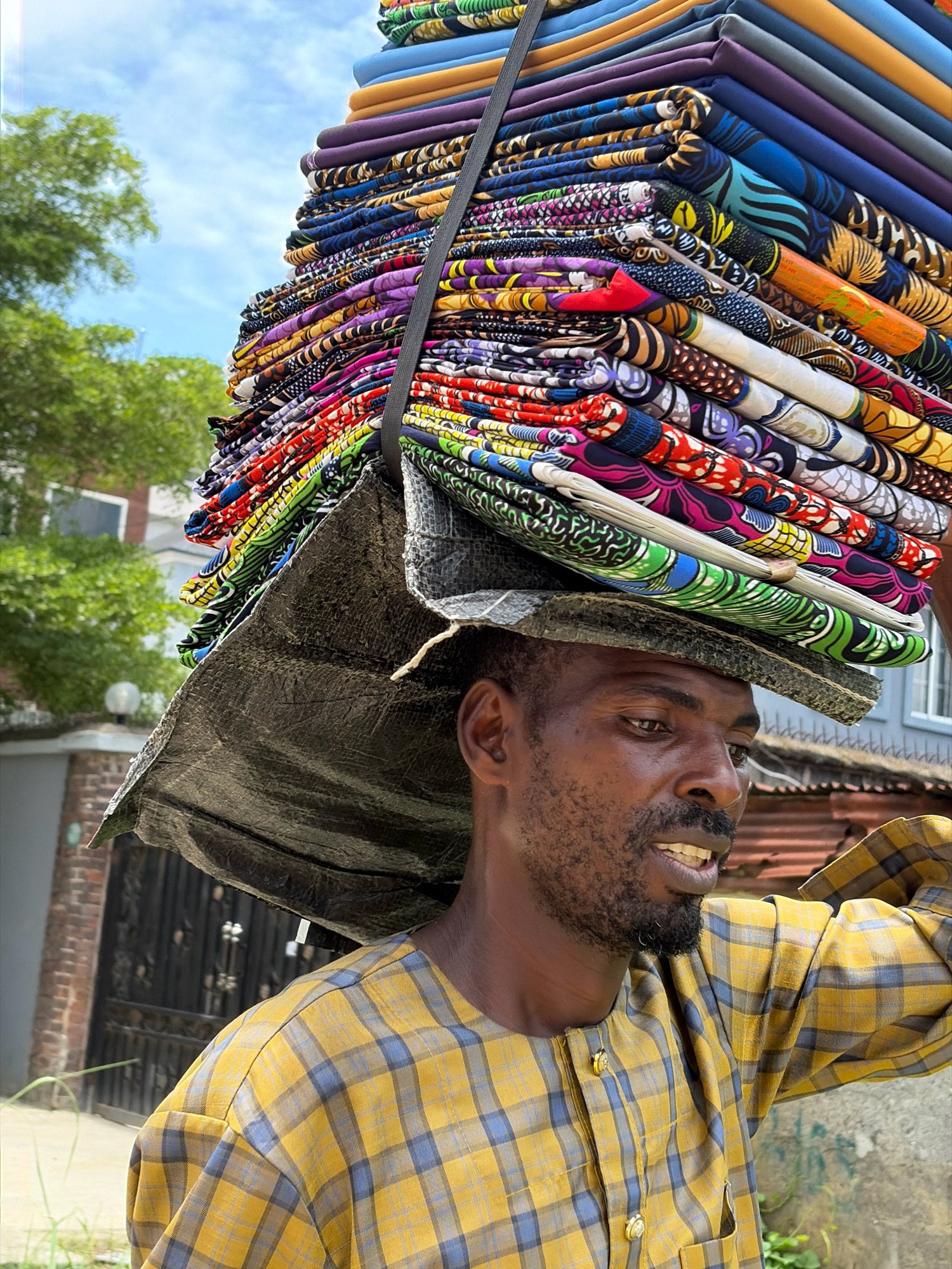 In Igboefon, Nigeria, a man carries a large stack of vibrantly patterned fabrics on his head, balanced with a tarp. He is wearing a yellow and blue plaid shirt. The man appears to be looking to his right, with his mouth slightly open. Behind him, lush green trees and residential buildings are visible under a bright, cloudy sky. The scene depicts a common sight of commerce and daily life in the area.
