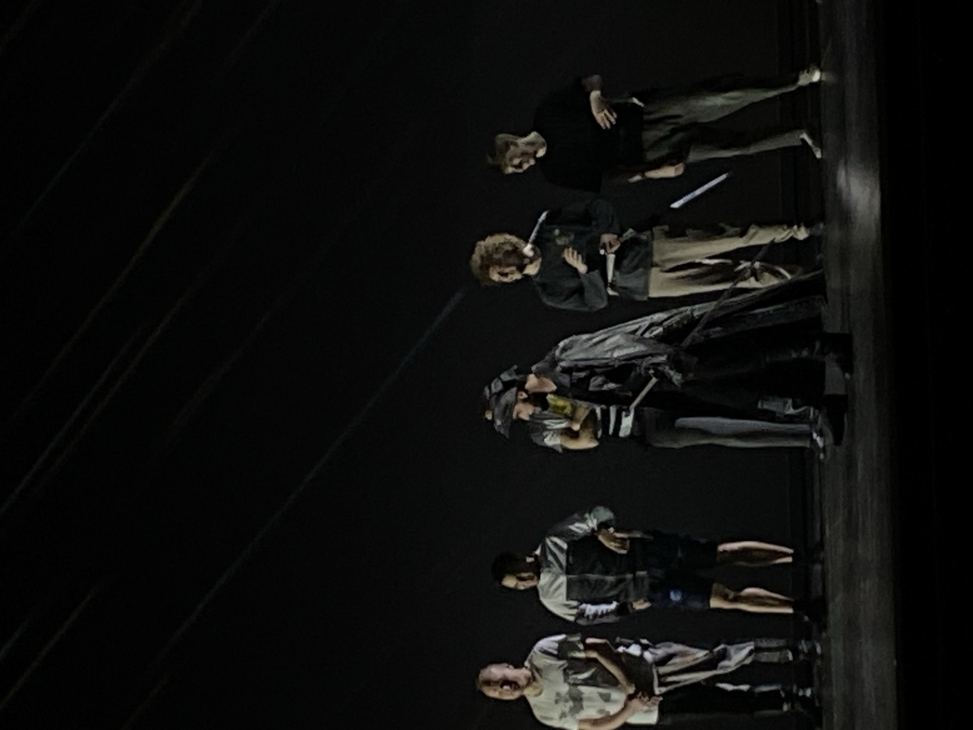 Five individuals are on a stage, presumably in Шебунино, Russia. The lighting is dim, casting a sense of anonymity on the scene. From top to bottom, the individuals are dressed distinctively: the first in a black shirt and tan pants, the second in a dark jacket with tan pants, the third in a dark coat, the fourth in a vest, and the last in a light shirt. They are positioned in a straight line, facing toward the viewer. Their postures vary. Some are relaxed, while others have their arms crossed or are looking away. The backdrop is dark and nondescript.