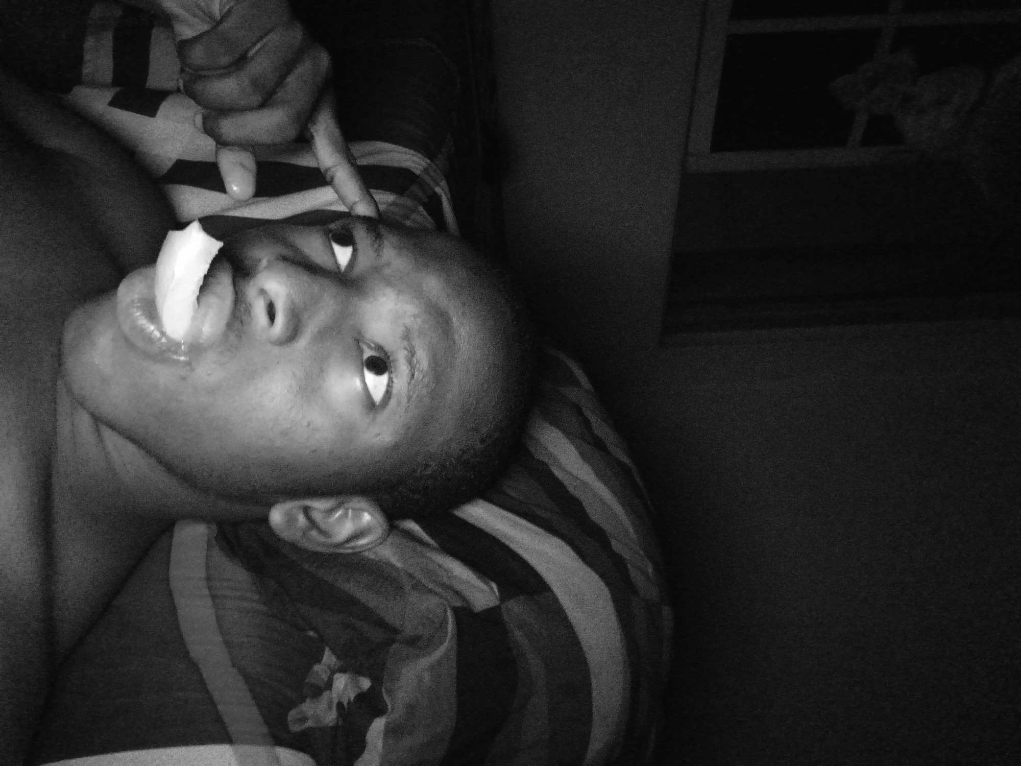 A black and white image shows a dark-skinned person lying on a striped surface, possibly a bed. They have a white object, possibly food, sticking out of their mouth. Another person's hand and finger are near their forehead. The location is suggested as Lagos, Nigeria, although there are no visual cues to confirm this. The environment is dark, with a window visible in the background. The lighting is poor, obscuring details.