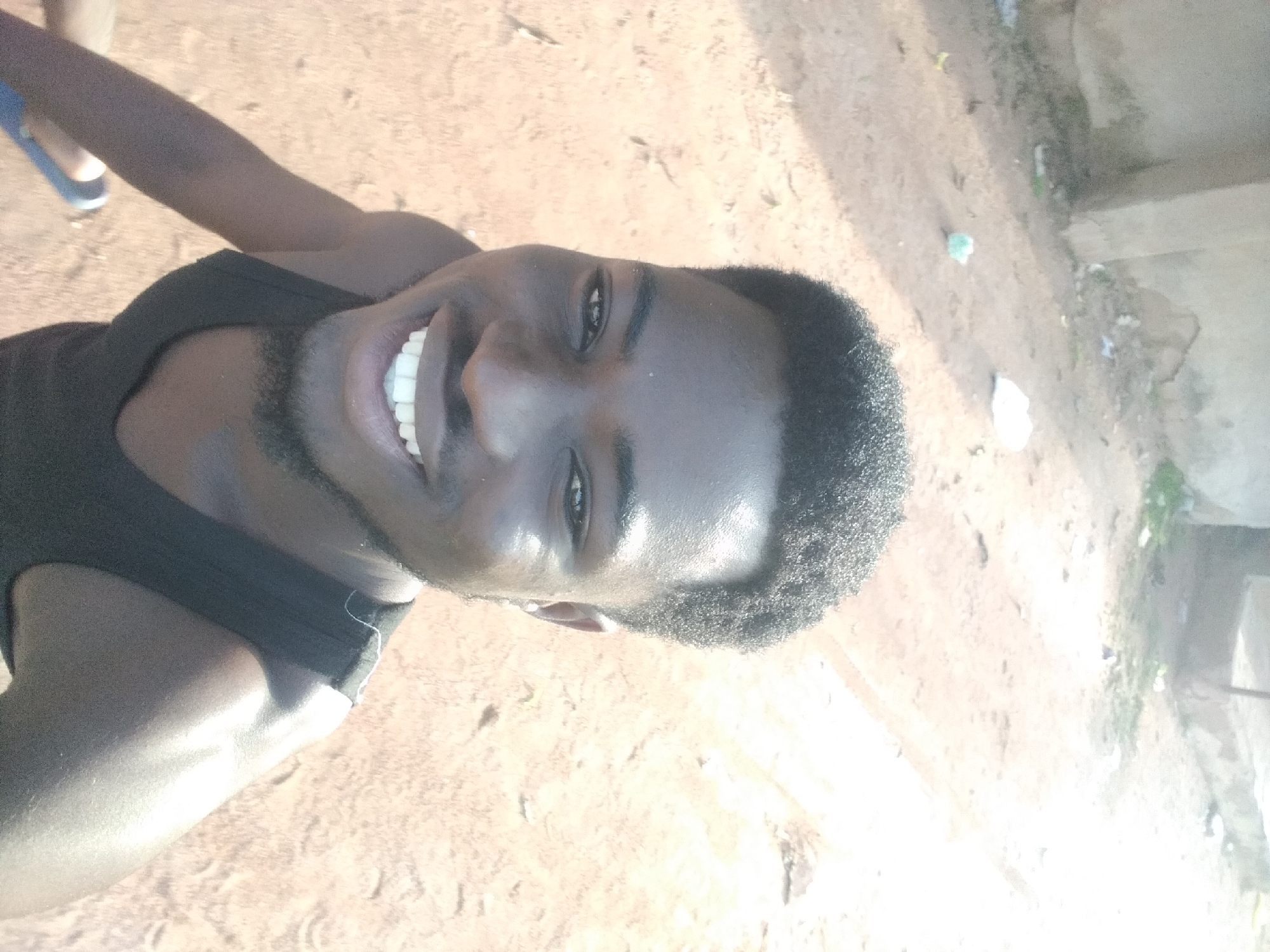 The image is a selfie taken outdoors in Jalingo, Nigeria. The subject is a young man with dark skin, short, curly black hair, and a short beard. He is wearing a black tank top and smiling. The background appears to be a dusty, outdoor area with a light-colored wall or building to the left and ground that is mostly dirt with some sparse vegetation. The lighting suggests it is daytime, possibly late afternoon due to the long shadows and bright sun. There is some scattered debris on the ground, including a white plastic bag. The overall impression is that of a casual, everyday scene in an urban or semi-urban environment.