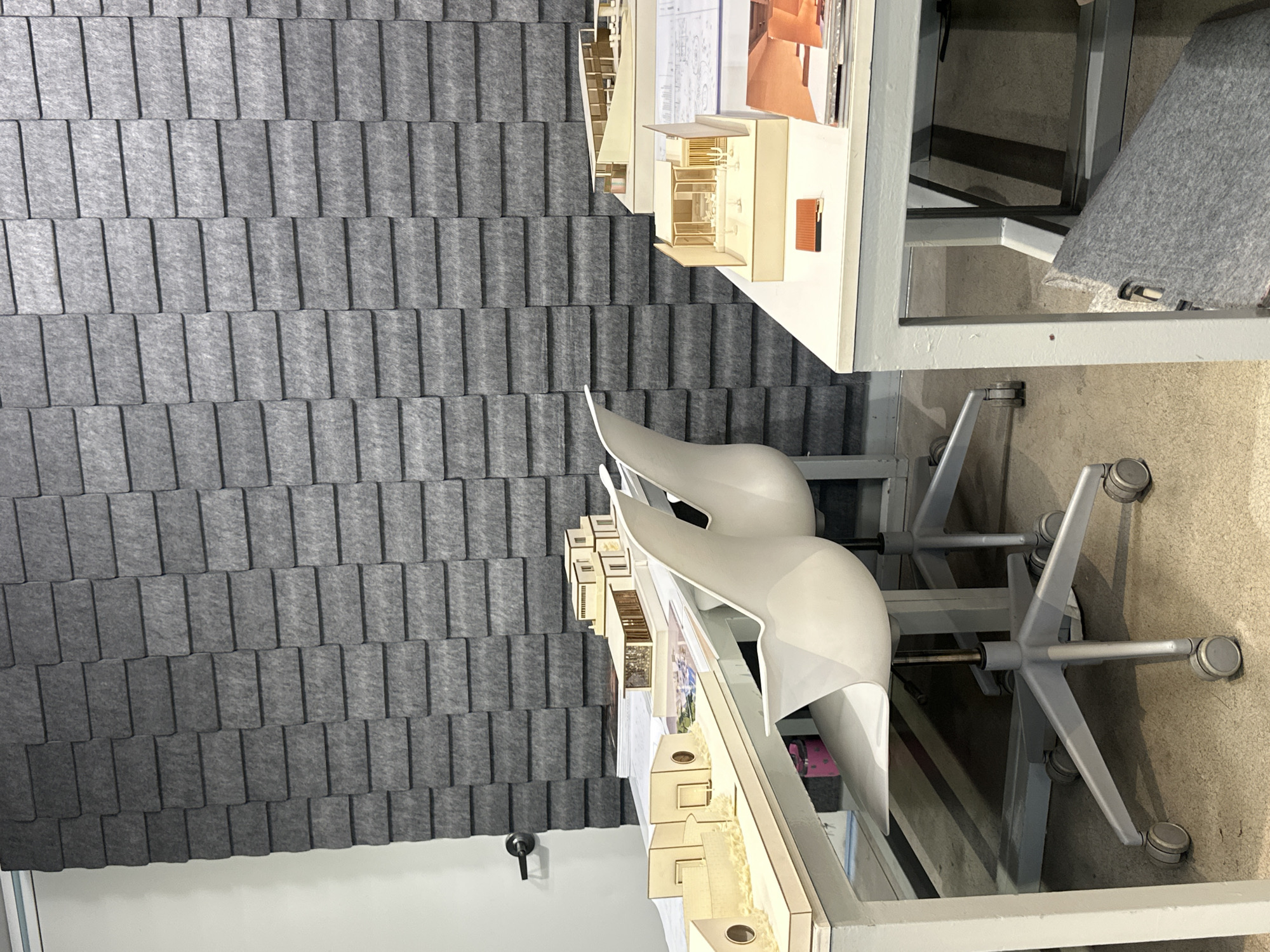 The image shows an indoor space with several elements. A gray, textured wall panel resembling staggered tiles is visible on the left. In the center, there's a modern white chair with a unique back design, along with a table holding architectural models and drawings. The models appear to be made of light-colored wood and cardboard. The table has a metal frame with a white top. The floor is a neutral color, possibly concrete. The room is well-lit, and the perspective suggests the photograph was taken at eye level. There are no people visible in the image. The location is unknown, and there's no definitive evidence to suggest it is in Mexico City, Mexico.