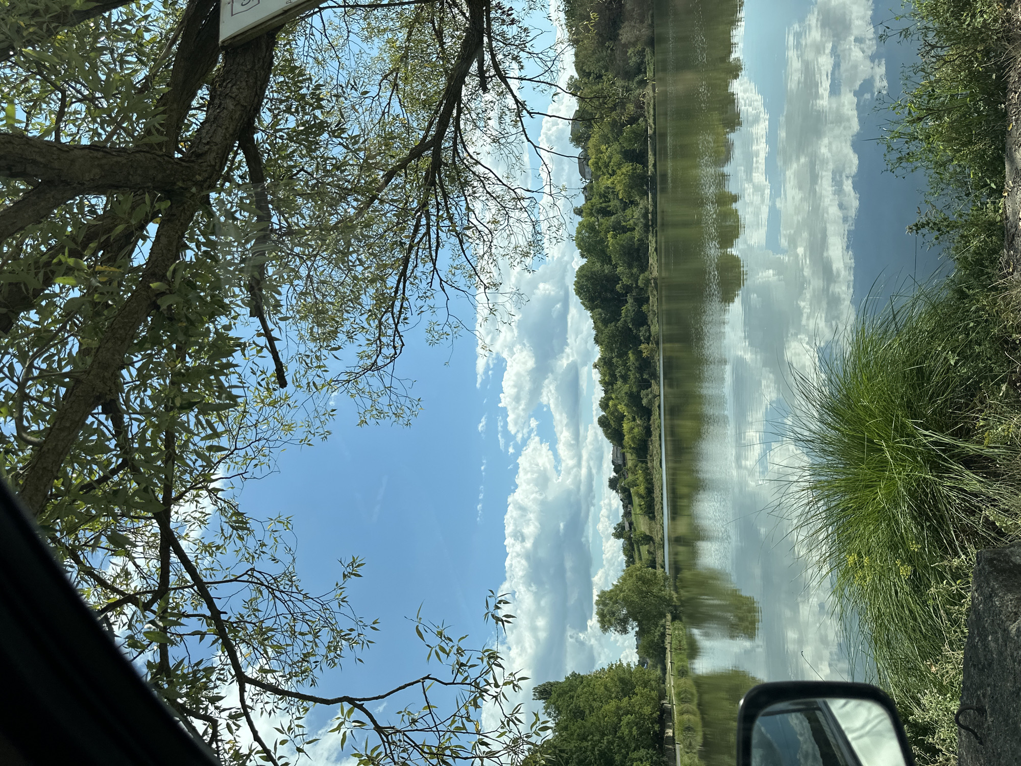 The image depicts a tranquil lake scene, likely in Makiv, Ukraine, on a clear day with scattered cumulus clouds. The foreground features the side mirror of a vehicle on the left, reflecting the sky and clouds. Lush green grass and vegetation grow along the shore. The lake itself is calm, with a near-perfect reflection of the sky, clouds, and the tree-lined horizon. The middle ground shows a dense line of green trees and some buildings in the distance, suggesting a rural or semi-rural setting. Overhanging branches of a large tree frame the top portion of the image, with dappled sunlight filtering through its green leaves. The overall impression is one of serene natural beauty.