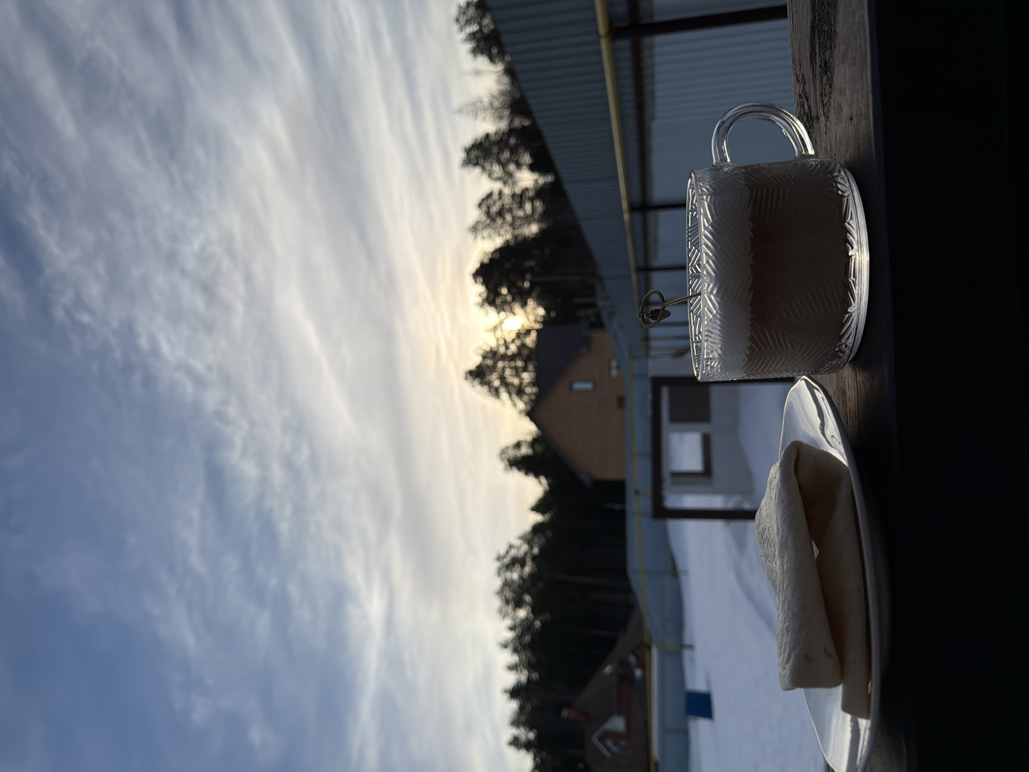 The image depicts an outdoor winter scene in Chaikovsky, Russia, during what appears to be either sunrise or sunset. In the immediate foreground, resting on a dark, textured surface, are a white oval plate containing two light-colored, folded items resembling tortillas or flatbread, positioned to the left. To the right, a clear glass mug with a geometric patterned exterior holds a light brown beverage topped with foam, into which a small, decorative metallic stirring tool or pick is inserted.
The midground features a wide expanse of snow-covered ground. A corrugated metal fence, likely galvanized or painted gray, spans horizontally across the scene, with a yellow gas pipe running parallel to it. Behind the fence, several houses are visible, including a prominent light brown brick-colored house with a dark roof to the right of center, and a smaller house with a reddish roof partially visible on the far left. A dense line of dark evergreen trees forms a treeline behind the structures.
The background shows a cloudy sky, transitioning from lighter blue at the top to a warm, soft glow near the horizon, where the sun is partially obscured by clouds and the treeline. No people or overt actions are depicted.