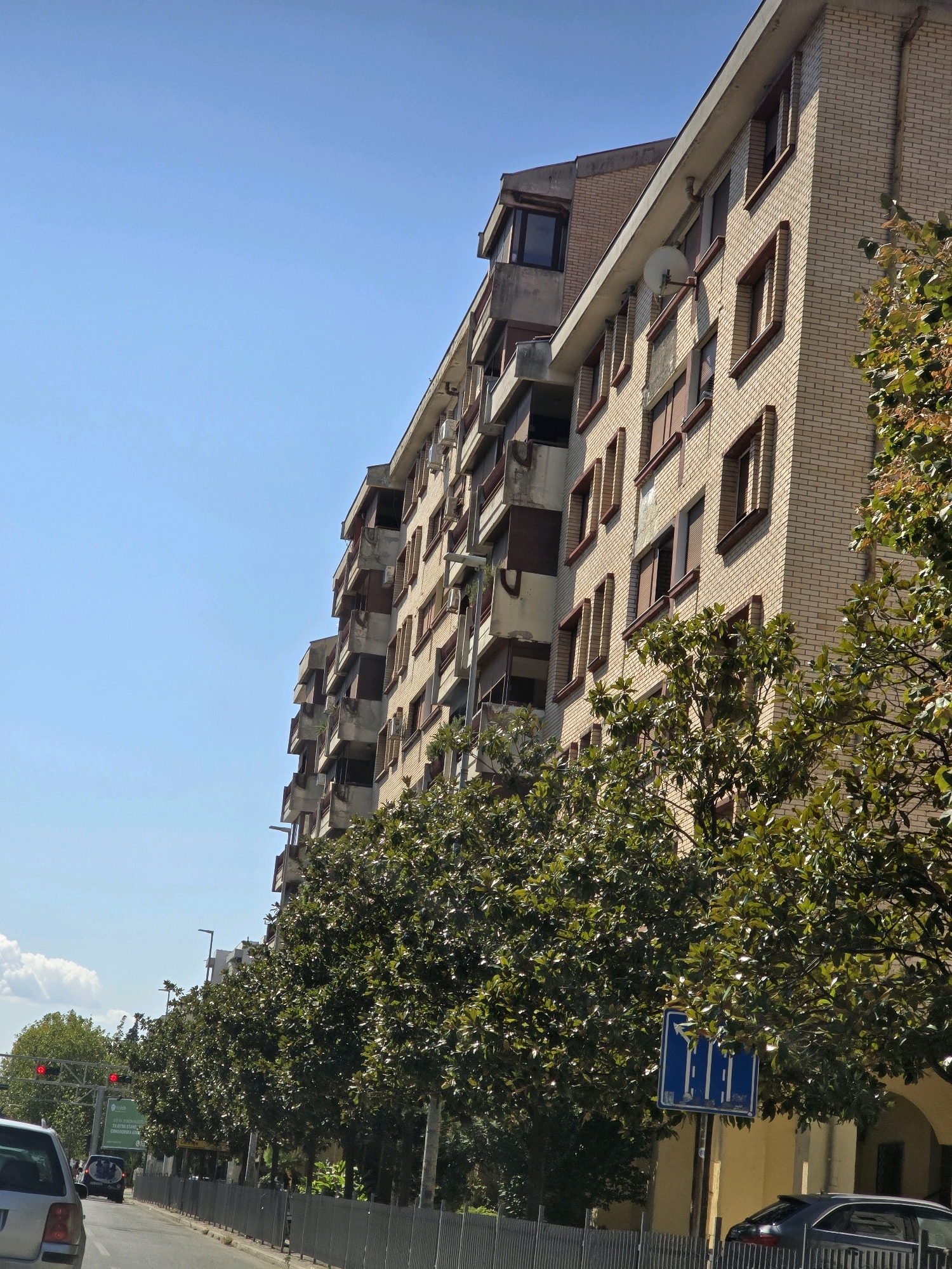 This is a medium-angle, eye-level, outdoor, daytime photograph taken in Podgorica, Montenegro. The image features the facade of a multi-story apartment building constructed with light-colored brick. The building has a series of balconies with dark-colored railings and windows with brown frames. Some windows appear to be covered by blinds or shutters. A satellite dish is visible on the exterior wall of the building. In the foreground, a row of mature trees with dark green foliage lines a sidewalk. Beyond the trees, a metal fence separates the sidewalk from a street where a gray car is visible. Traffic lights, a street sign with blue background and white arrows, and another green traffic sign are also present. The sky is clear and blue, with a few scattered white clouds on the left side of the frame. The architecture suggests an urban environment, likely a residential area within Podgorica.