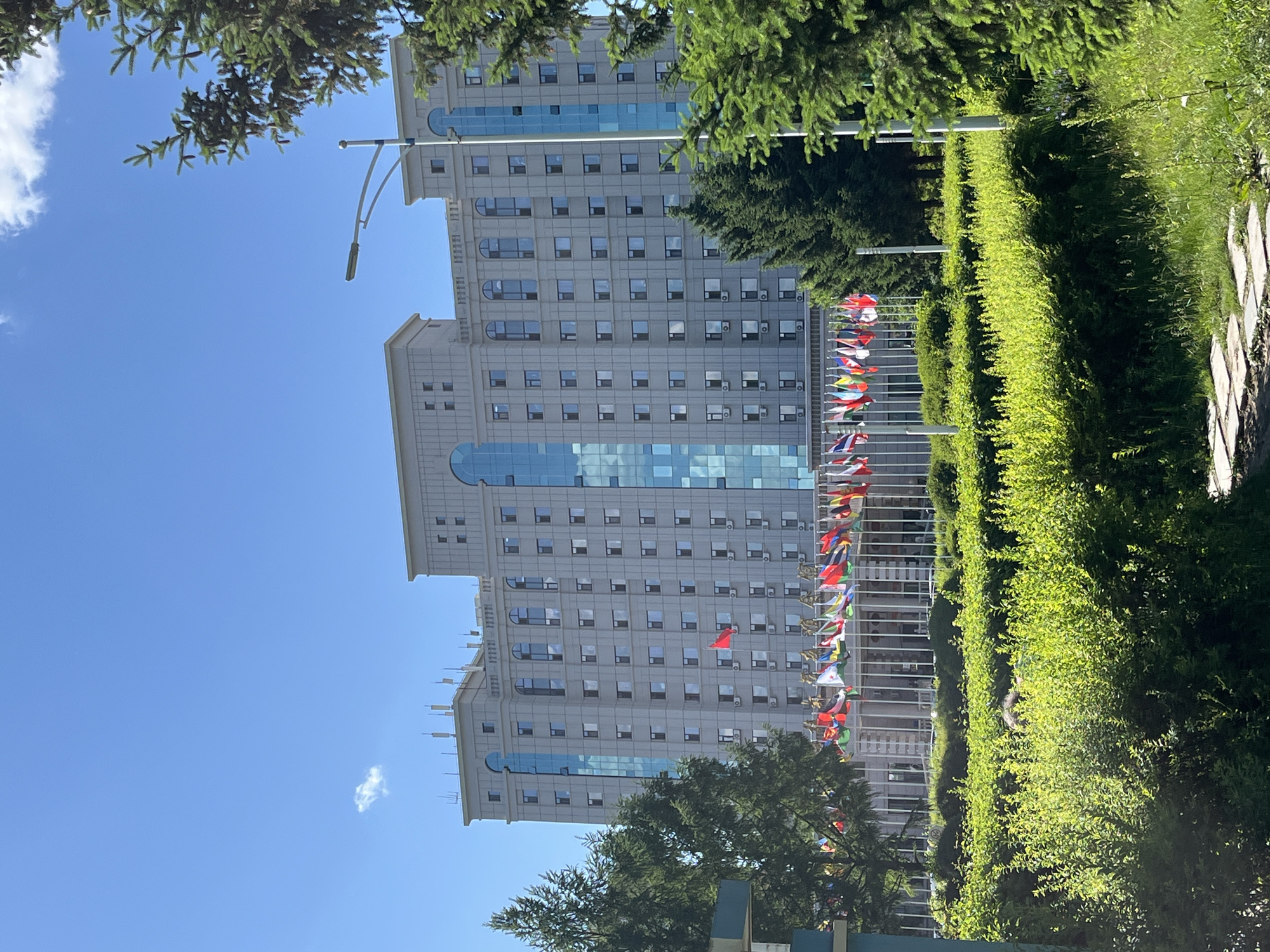 The image depicts a multi-story building in Changchun City, China. The structure is rectangular, light gray, and features rows of windows. A central glass section runs vertically through the building's facade. In front of the building, a row of flagpoles displays various national flags. Lush green hedges flank a sidewalk, partially obscuring the view. The sky is clear blue, with a few scattered clouds. Trees are visible in the foreground, framing the scene.