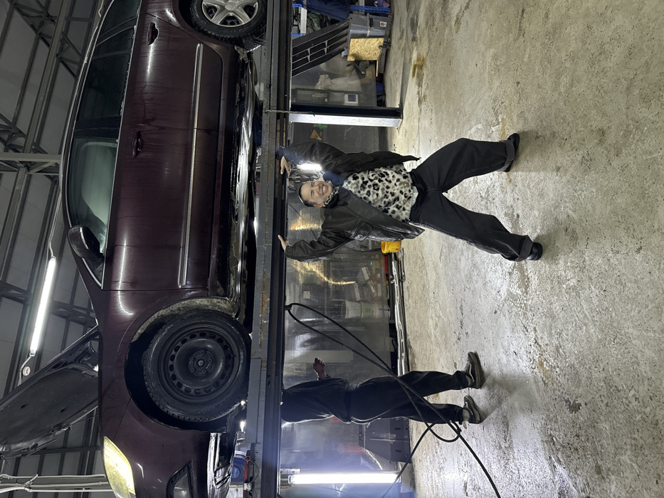 A maroon car is lifted on a hydraulic lift in an auto shop. A person in a leather jacket and animal print sweater stands beneath the car, holding onto the lift arms and smiling. Another person stands further back, holding a cord, likely a mechanic. Fluorescent lights illuminate the scene. The floor is concrete.