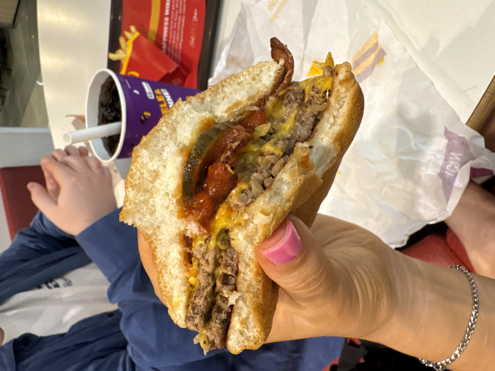 A person with pink nail polish holds a halved double cheeseburger. Visible burger ingredients include two beef patties, melted cheese, bacon, pickles, and ketchup. A McDonald's french fry container and a McDonald's drink cup with a straw are in the background. The location is Spreitenbach, Switzerland, identifiable by the McDonald's packaging. In the bottom left corner, a child's hand rests on a red surface. A person's leg and a silver chain bracelet on their wrist are visible in the lower right. Crumpled fast-food wrapping paper is in the background near the burger.