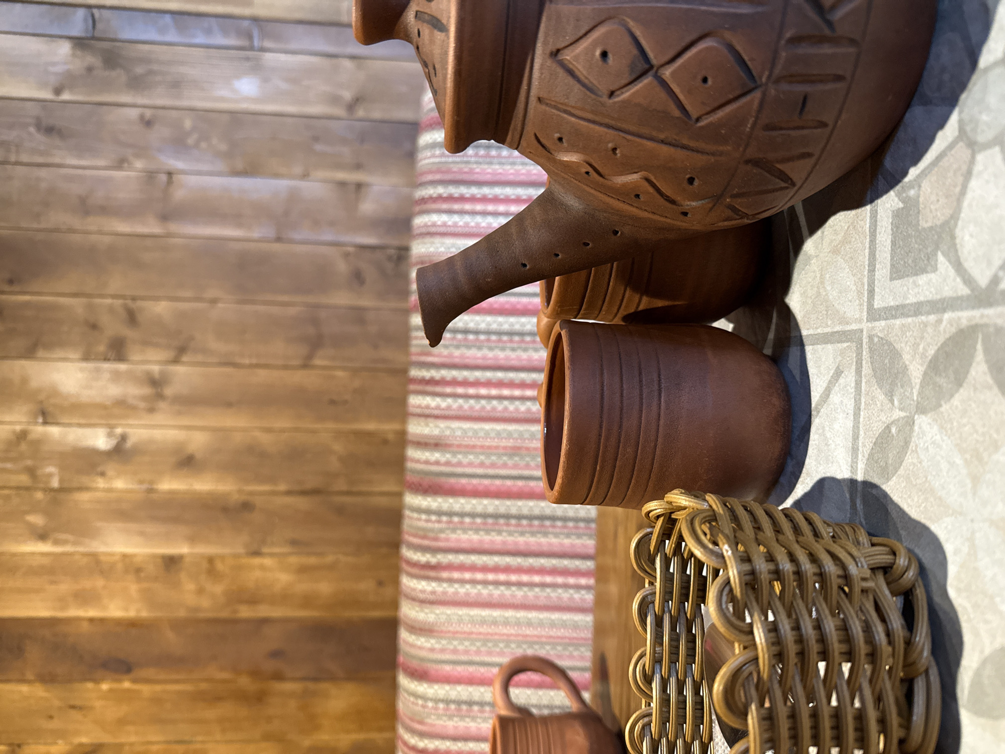 This is a close-up, eye-level shot of a table setting in what appears to be a cafe or restaurant. The background features a vertical wooden wall paneling. In the mid-ground, a padded bench with a striped upholstery of pink, white, and gray is visible. The foreground is dominated by a table with a patterned surface, featuring ceramic mugs and a teapot.
Specifically, on the table, there is a woven basket in the lower left corner, appearing to be made of light brown plastic or wicker. To the right of the basket are two unglazed, reddish-brown ceramic cups with ribbed detailing. Further to the right, a larger, matching reddish-brown ceramic teapot is prominently displayed. This teapot has an angular spout and decorative etched patterns resembling diamonds and zigzag lines, along with small perforations on the spout. Another unglazed ceramic cup is visible behind the teapot.
The lighting suggests an indoor setting, possibly with natural light supplemented by artificial light sources, casting soft shadows. The overall ambiance is rustic and cozy. The image was taken in Izhevsk, Russia.
