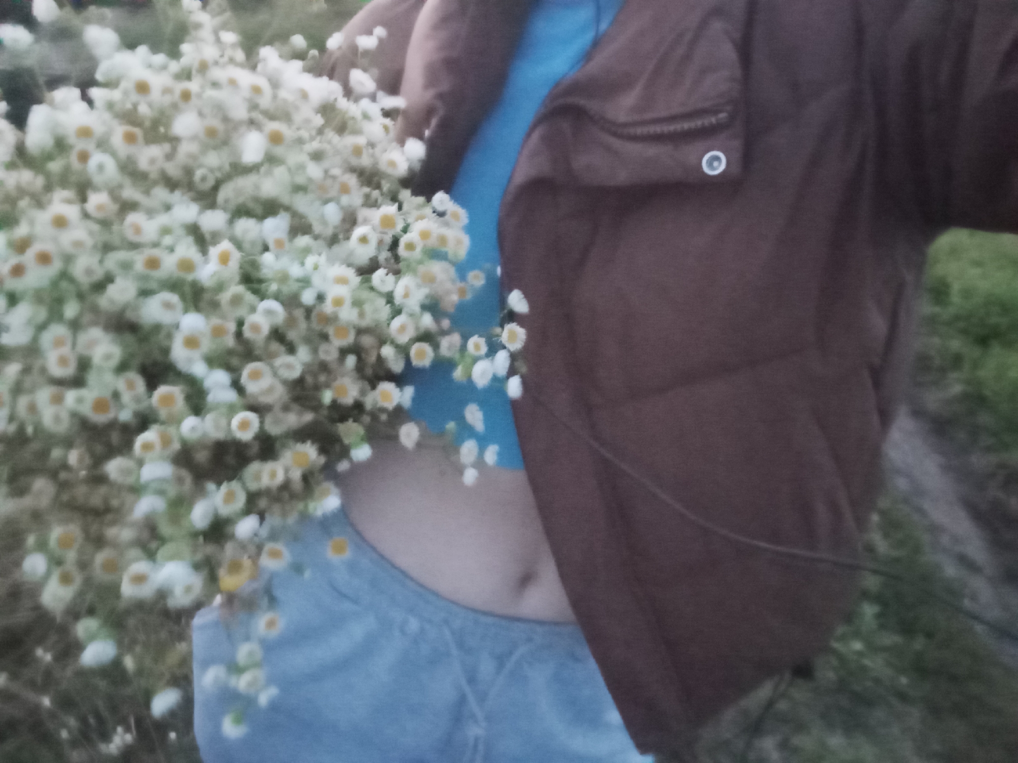 A person wearing a brown puffer jacket, a blue t-shirt, and gray sweatpants is holding a large bouquet of small white and yellow flowers. The person's midriff is visible. The background consists of green grass. The image appears to be taken outdoors.