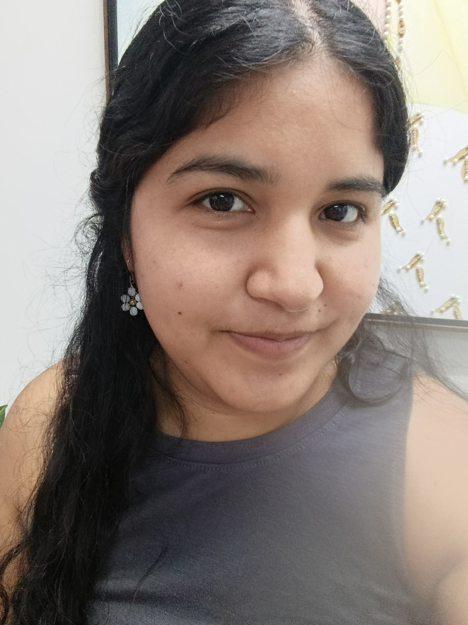 The original image is a medium shot of a young woman with dark, wavy hair. She is wearing a dark gray tank top. She has a small, flower-shaped earring with light blue and clear beads on her left ear. Her skin is light brown and she has dark eyes and eyebrows. She is looking slightly to her right and smiling faintly. The background appears to be a wall with some decorative elements on it, possibly a frame and some golden, shrimp-shaped ornaments. The scene is lit by artificial light.
The provided crops focus on different parts of the woman's face and upper body, highlighting her eye, hair, earring, and the texture of her shirt and skin. The crops do not offer any additional environmental context that would help determine the specific location within Juan Griego, Venezuela.