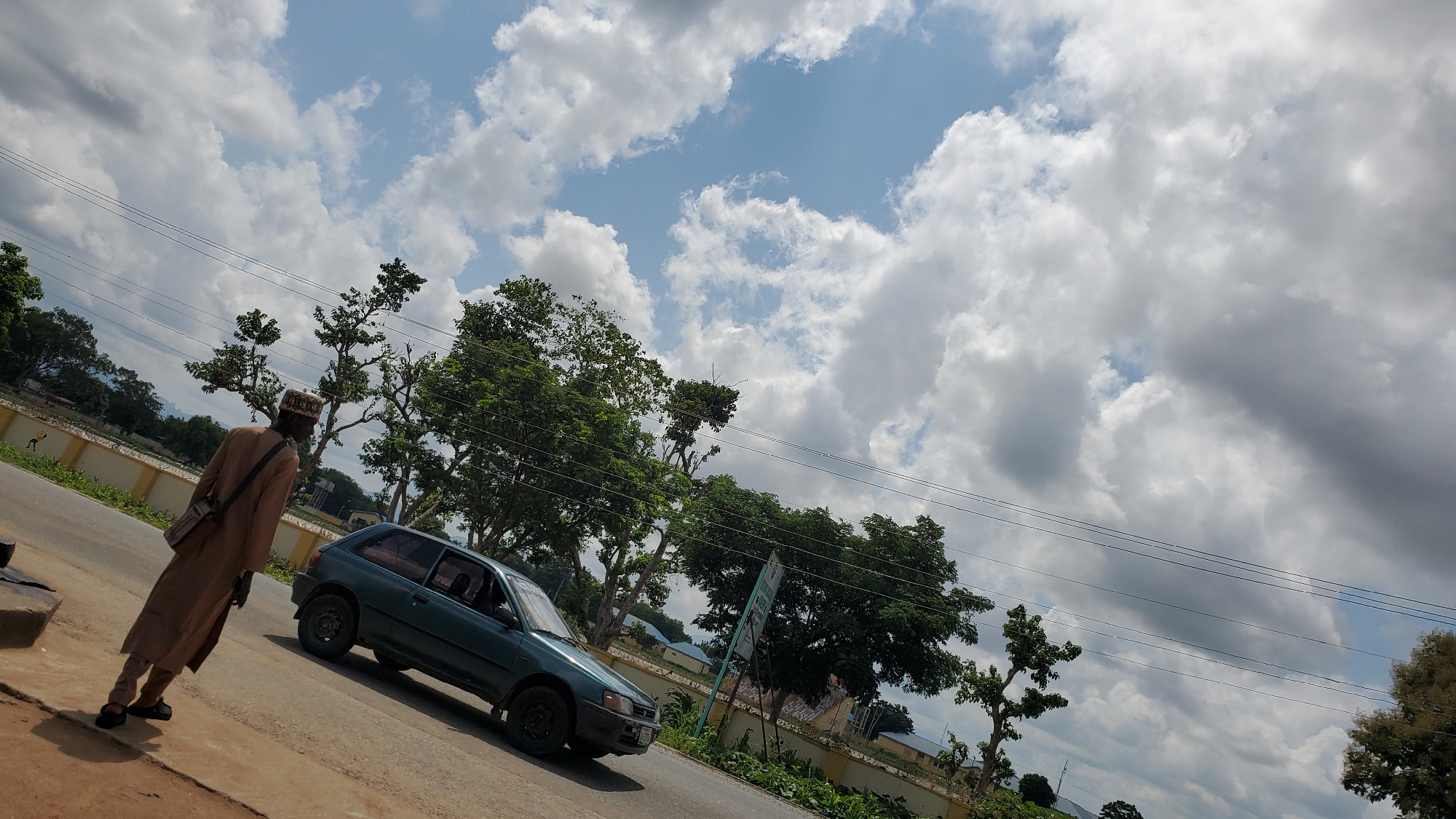 A lone man in traditional tan clothing and a patterned cap stands on the side of a paved road in Jalingo, Nigeria. He has a satchel slung across his right shoulder and is looking away from the camera. To his left, a dark green hatchback car is parked on the roadside. In the background, lush green trees line the road, and beyond them, buildings and more trees are visible. Overhead, a partly cloudy sky with large white cumulus clouds dominates the scene, with some power lines stretching across the frame. The ground in the foreground is a mix of reddish-brown earth and grey pavement. The overall impression is a snapshot of everyday life in a Nigerian city under a vast sky.