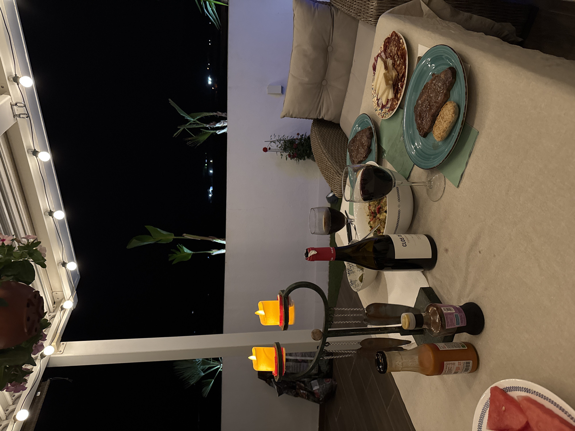 The image depicts a dimly lit outdoor dining setup on a terrace at night. In the foreground, a table is set with a light-colored tablecloth. On the table, there are two plates of food, each containing a steak and a small side. One plate also has a biscuit. There is a bowl of what appears to be a salad or pasta dish, a bottle of red wine, and two glasses of red wine. Two small bottles of condiments are also present. A decorative candelabra with two lit LED candles is to the left of the table. In the background, a white wall and some plants are visible. String lights are overhead, illuminating the scene. The furniture includes a cushioned chair or sofa to the right. The overall impression is of an intimate evening meal, likely in a residential setting. The location is specified as the city of Vallada, Spain.