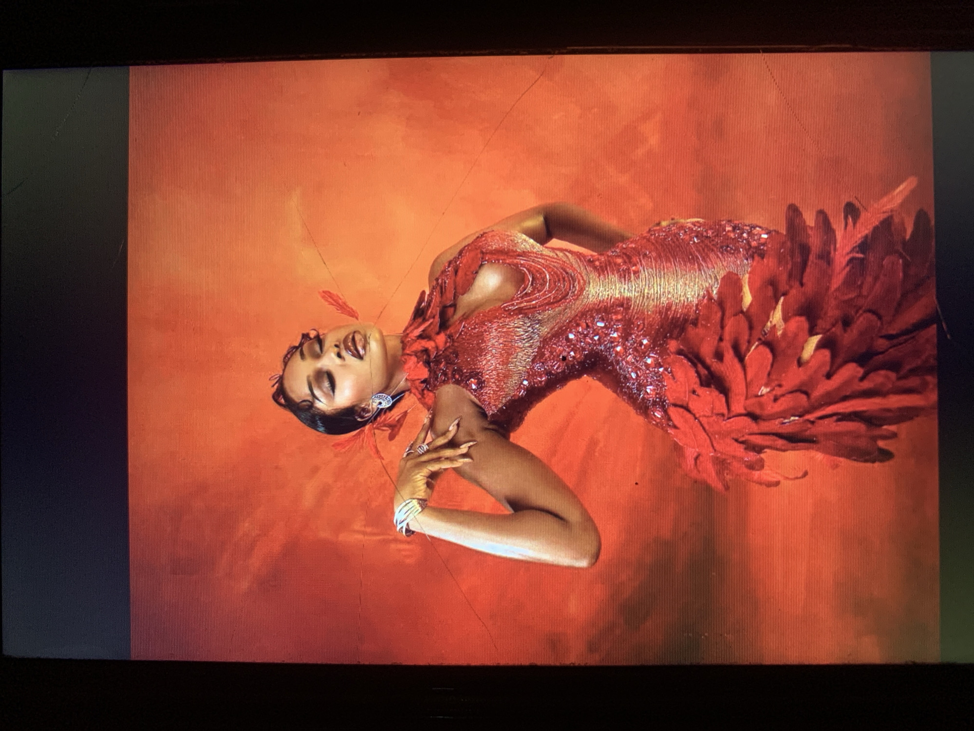 A woman in a red, feather-adorned dress is positioned against a red background. The image appears to be a photograph of a screen. The location mentioned, New Stadium, Nigeria, does not seem directly related to the visual content.