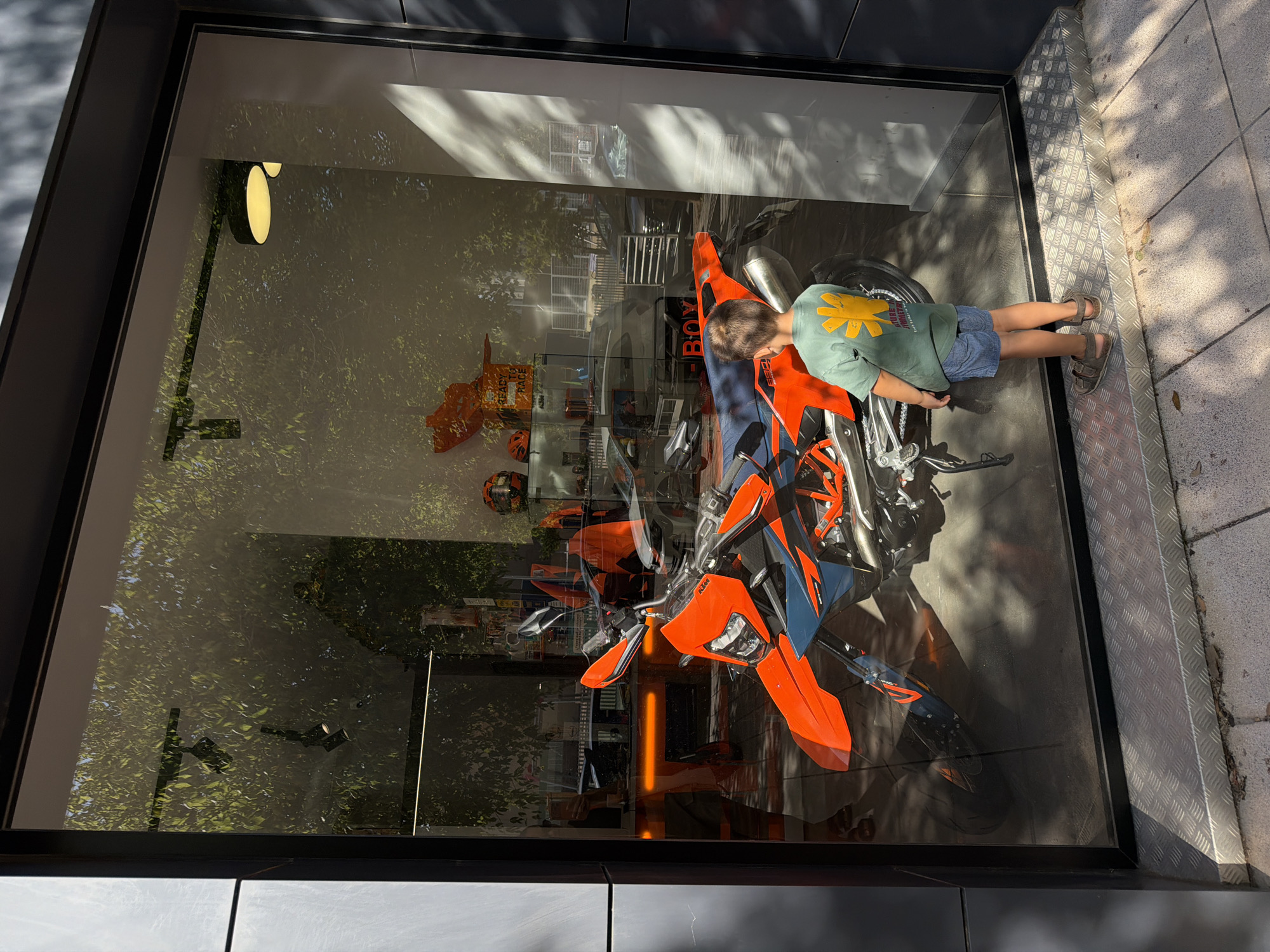 A young boy, seen from behind, wearing a green t-shirt with a yellow sun graphic and denim shorts, stands on a diamond-plate metal ramp looking into a store window. The boy is positioned to the right of the frame. Through the glass, an orange and blue dirt bike is displayed prominently. Behind the motorcycle, the store interior is visible, with shelving units displaying helmets, apparel, and other merchandise. Reflections on the glass show trees and buildings from the street outside, as well as internal lighting fixtures. The store appears to be a motorcycle dealership. The scene is outdoors, with sunlight casting shadows on the pavement and the metal ramp.